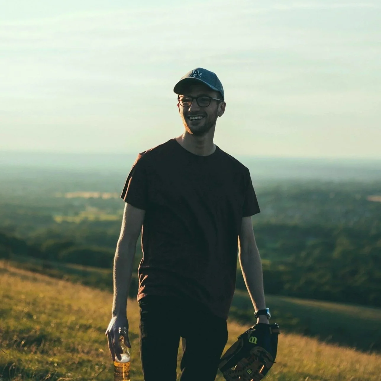 A young man wearing glasses and a baseball cap, smiling, standing outdoors in a field during sunset, holding a water bottle in one hand and a baseball glove in the other.