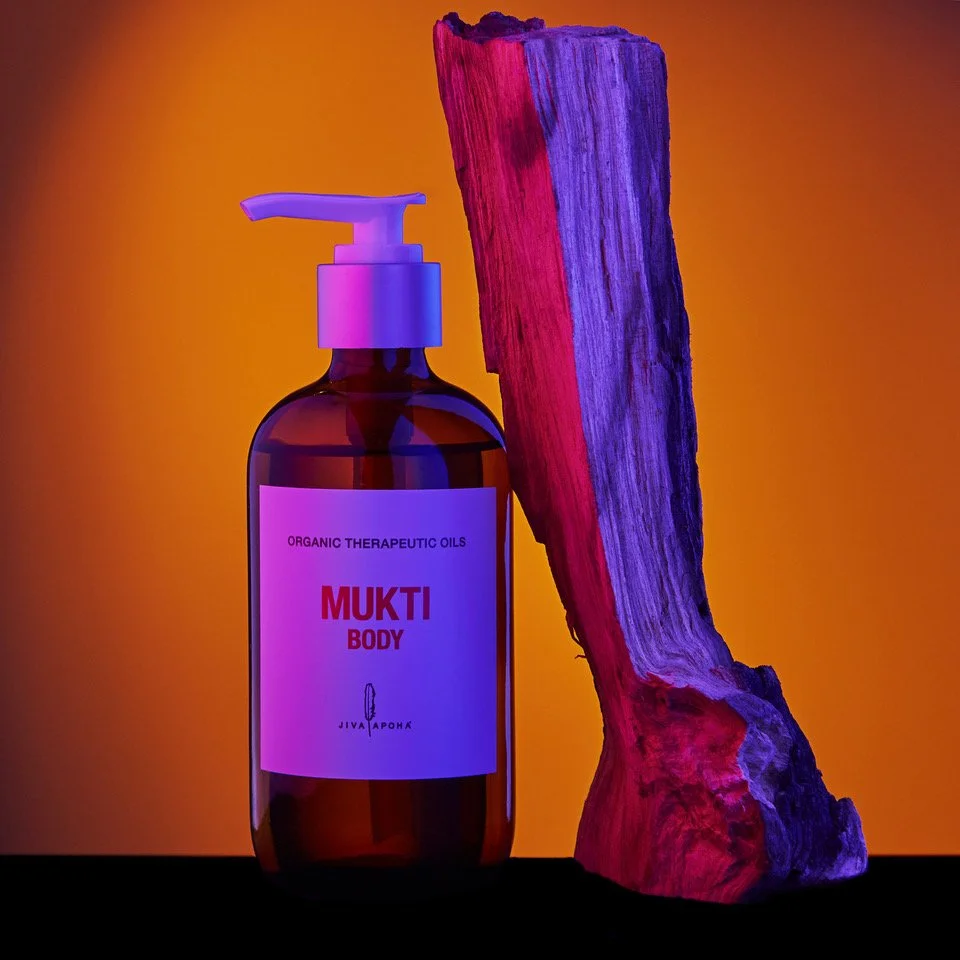 A bottle of organic therapeutic oils labeled 'Mukti Body' with a body pump, standing next to a large piece of wood painted with pink and purple lights, against an orange background.