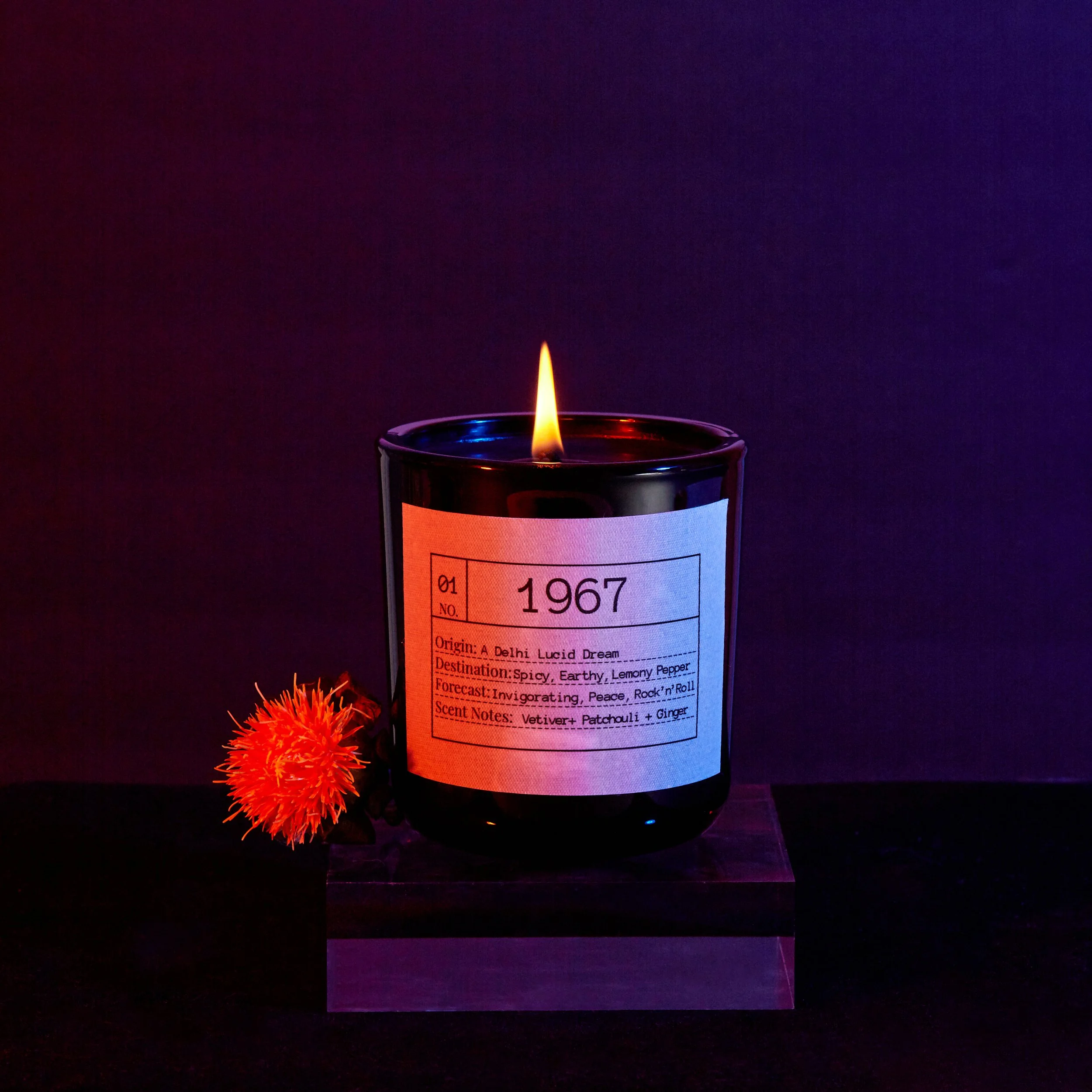 A burning candle in a black container with a label indicating a 1967 production date, along with a vibrant orange flower placed in front of it.