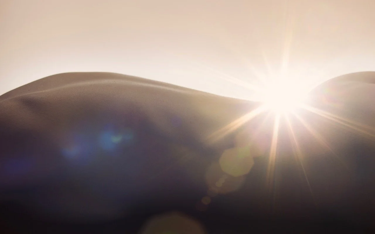 Sand dunes with the sun setting or rising behind them, creating a lens flare and bright light.