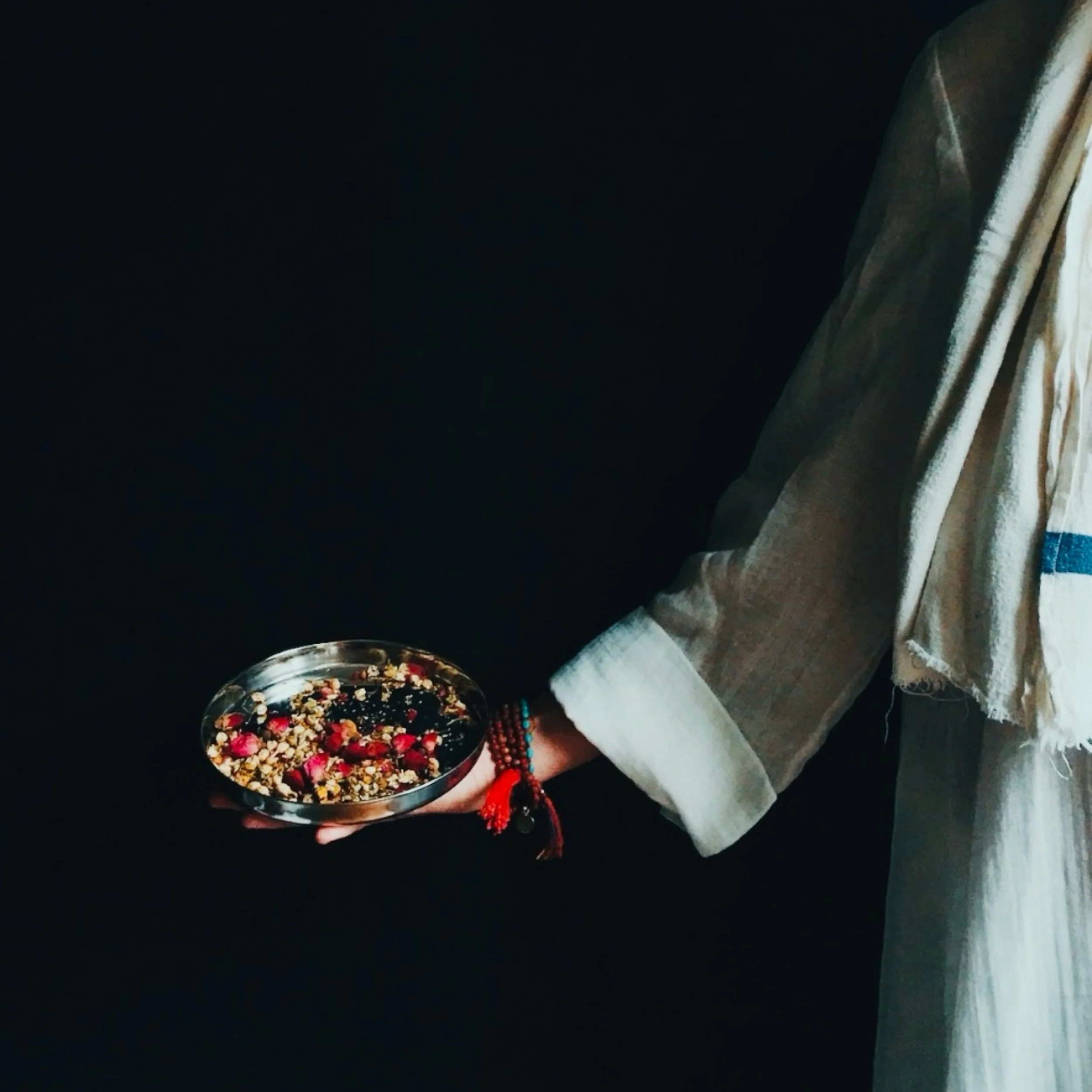 Person holding a bowl of mixed dried flowers, jewelry, and beads, wearing jeans and a white top, standing on a wooden floor.