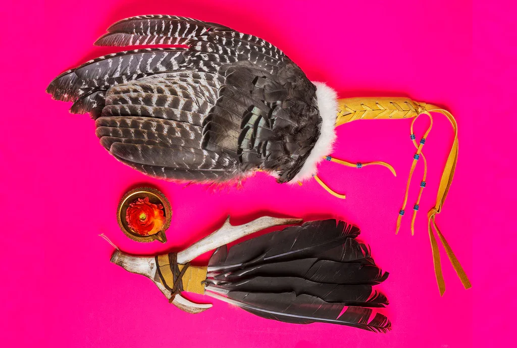 Decorative Native American headdress with feathers, a small bowl of red and orange flowers, and a feathered accessory on a pink background.