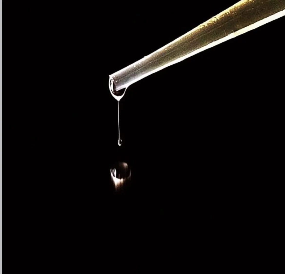 Close-up of a glass pipette with a drop of liquid falling from its tip against a black background.