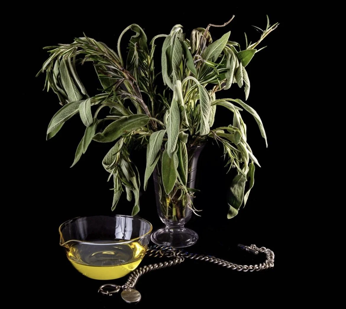 A glass vase with fresh herbs, a glass bowl with oil, and a silver necklace set against a black background.