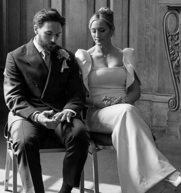 A black and white photo of a bride and groom sitting side by side, looking down with solemn expressions, against a wood-paneled background.