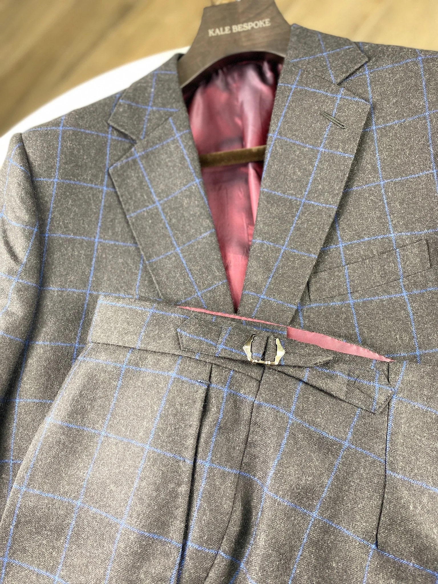 A close-up of a men's gray suit jacket with a blue windowpane pattern, displayed on a hanger with a pink lining inside.