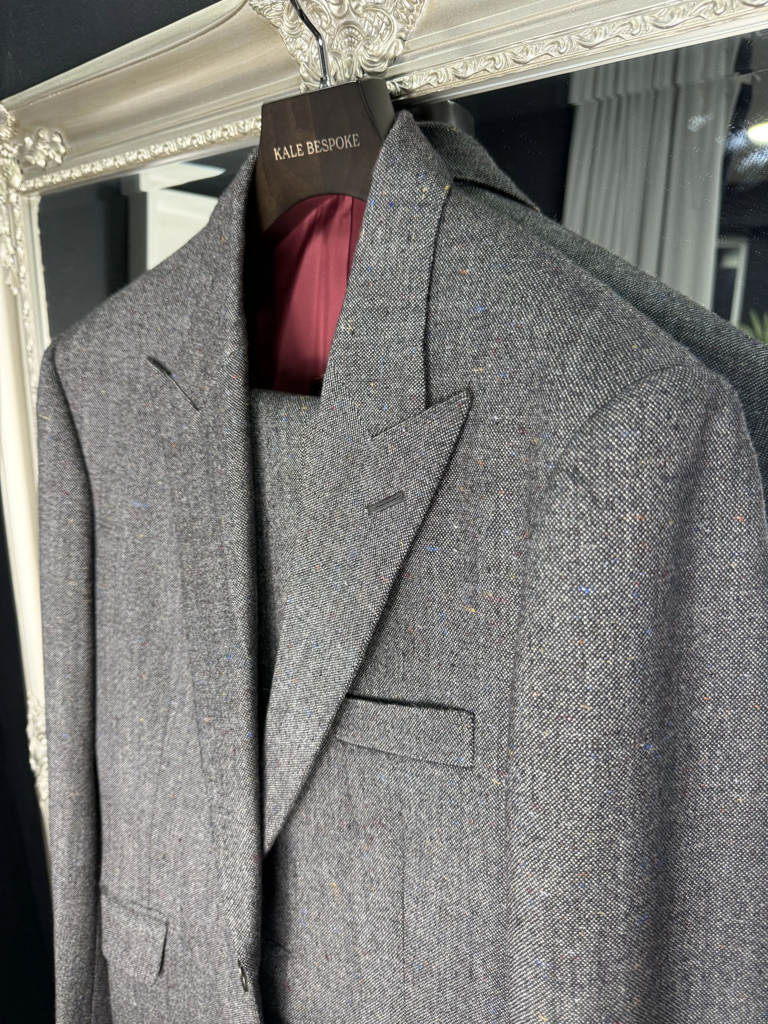 Gray tweed suit jacket hanging on a black hanger with a red interior. The hanger is labeled 'KALE BESPOKE'. The suit is displayed on a mirror with ornate silver detailing.