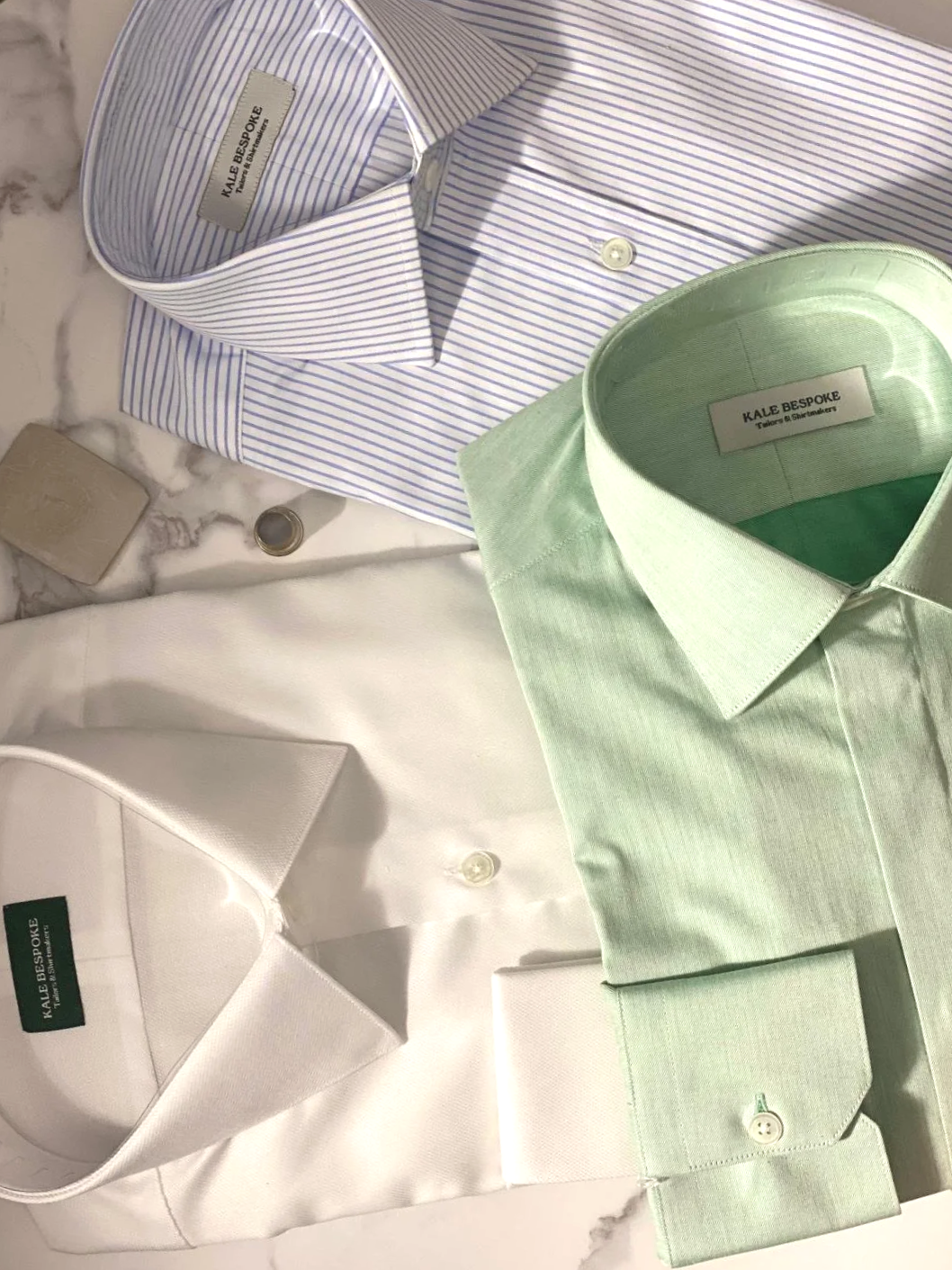 Three designer dress shirts in white, light green, and white with blue stripes, laid out on a marble surface. Each shirt has a label that reads 'KALE BESPOKE'.
