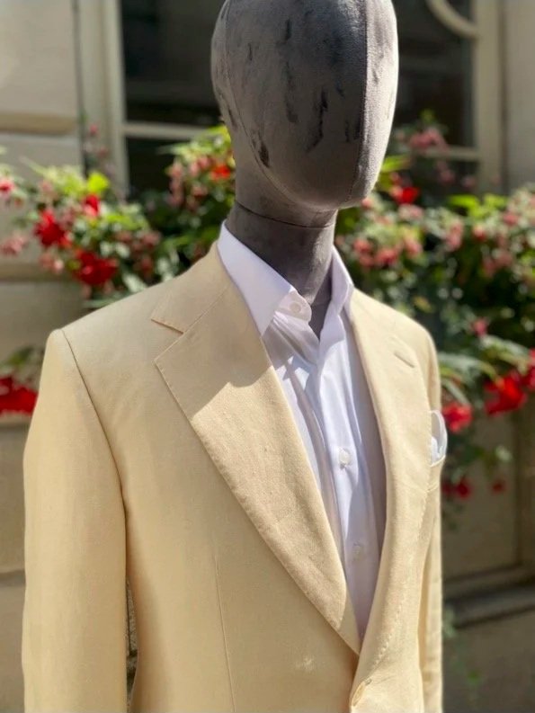 A mannequin dressed in a light cream-colored suit with a white dress shirt, positioned outdoors with flowers in the background.