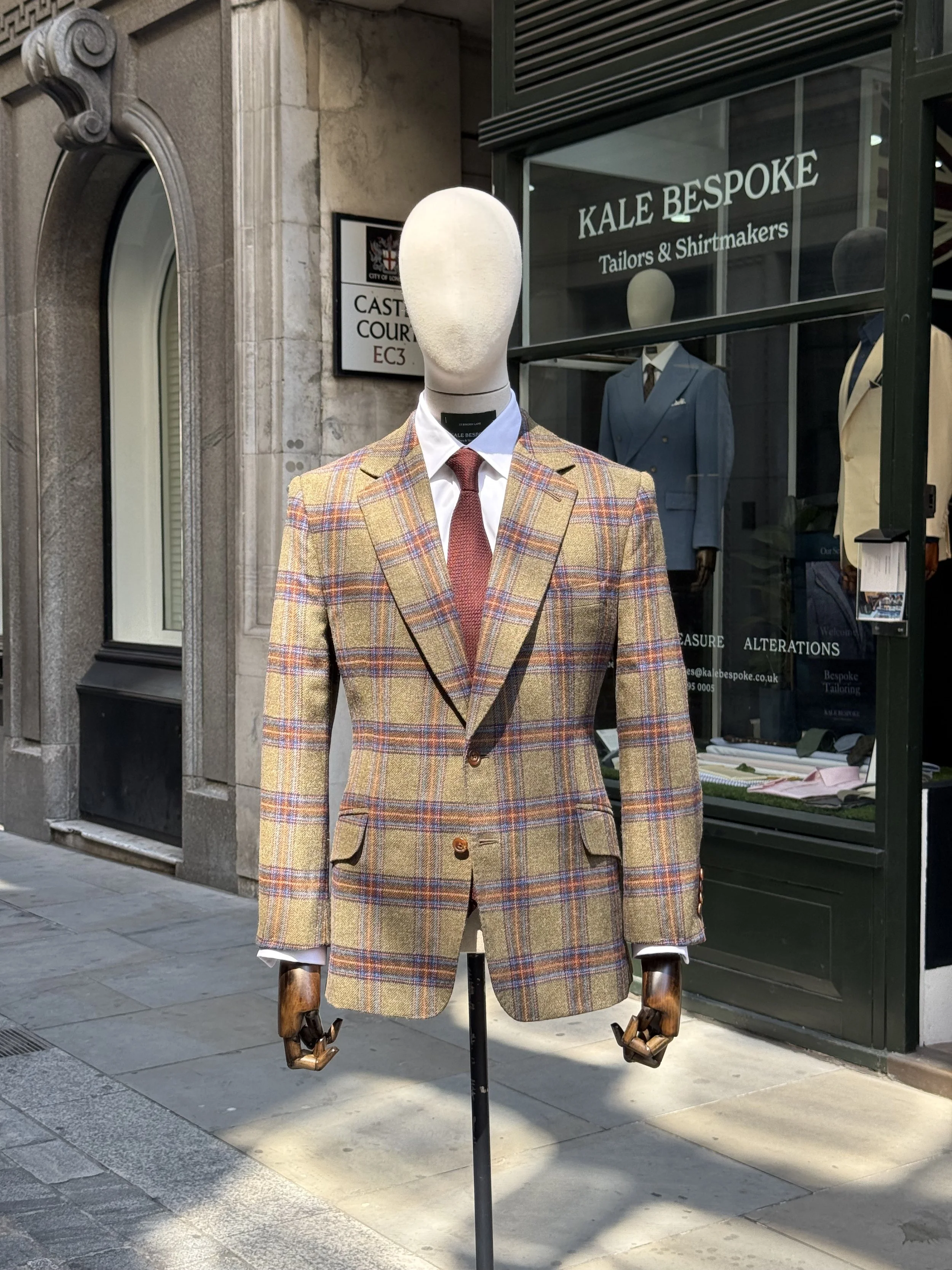 A mannequin dressed in a rally tweed fox flannel blazer, white shirt, and red grenadine tie, positioned outside a clothing store with a sign for Kale Bespoke Tailors &Shirtmakers, on  Birchin lane.