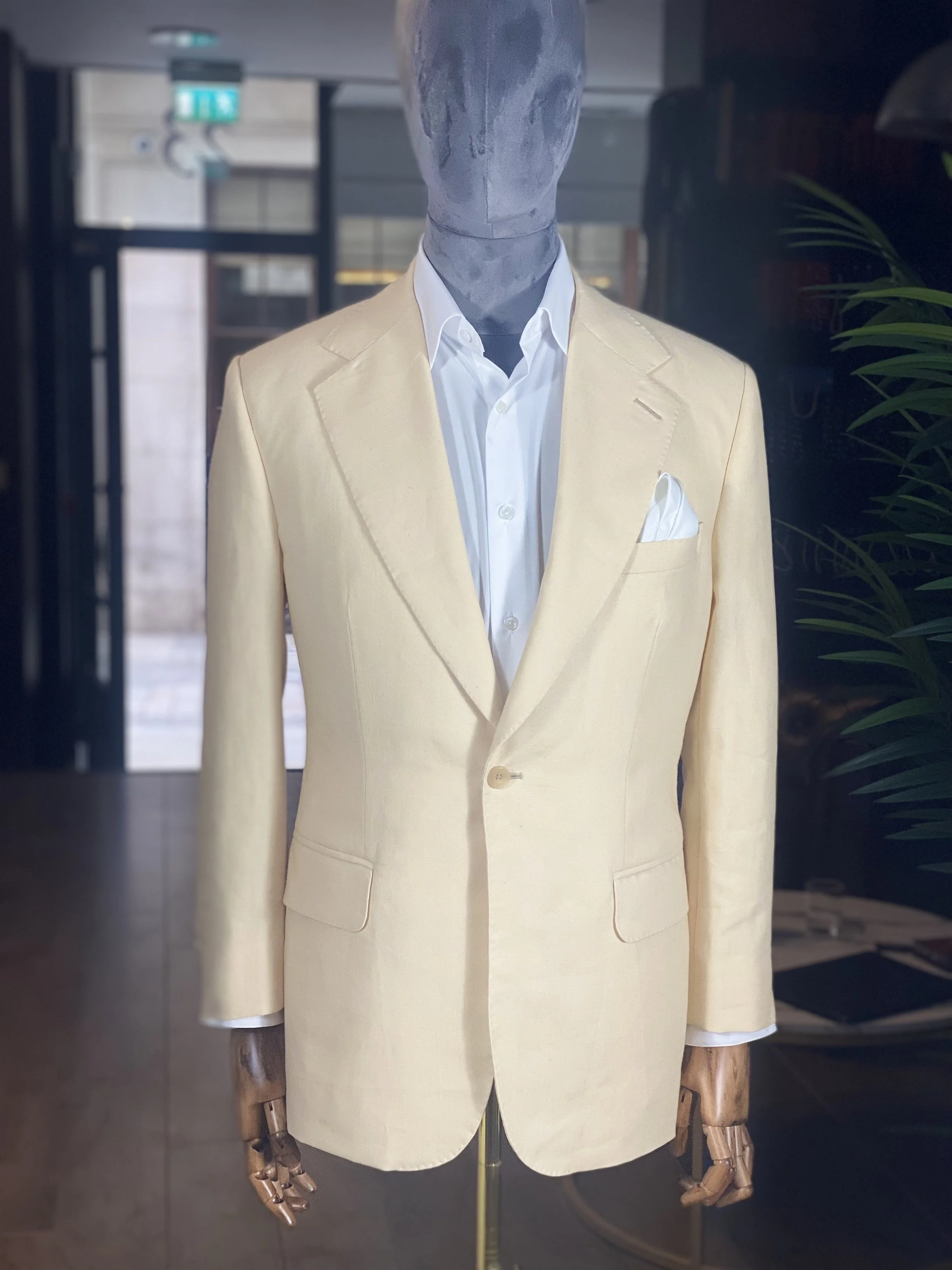 A mannequin dressed in a cream-colored blazer over a white shirt with a pocket square, standing indoors in front of a glass window or door.