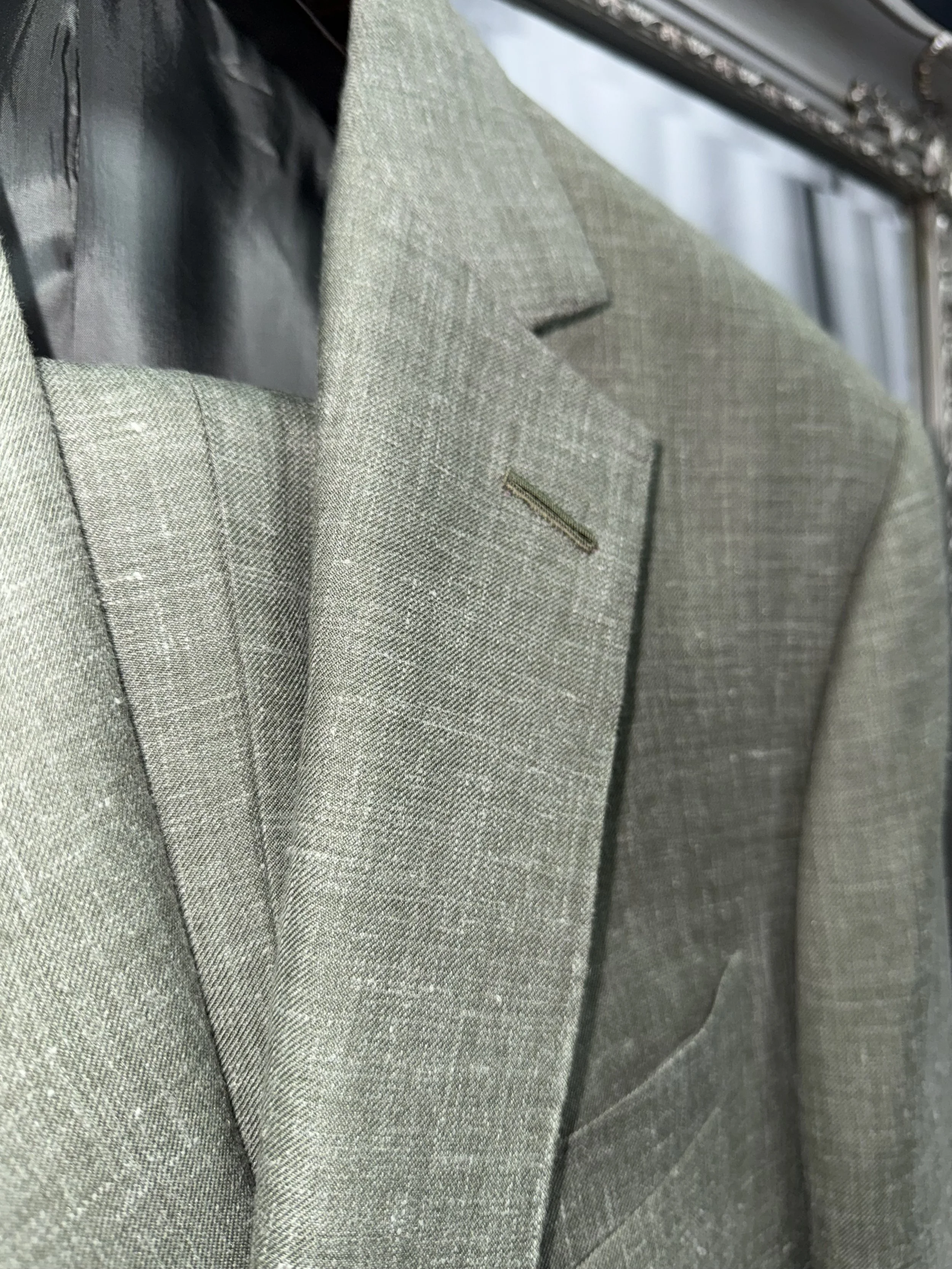 Close Up of a sage green wool, silk, linen blend  bespoke jacket featuring a notch lapel with hand finished Milanese buttonhole, softly structured shoulder and natural texture. Handmade bespoke tailoring displayed on hanger inside our shop on Birchin