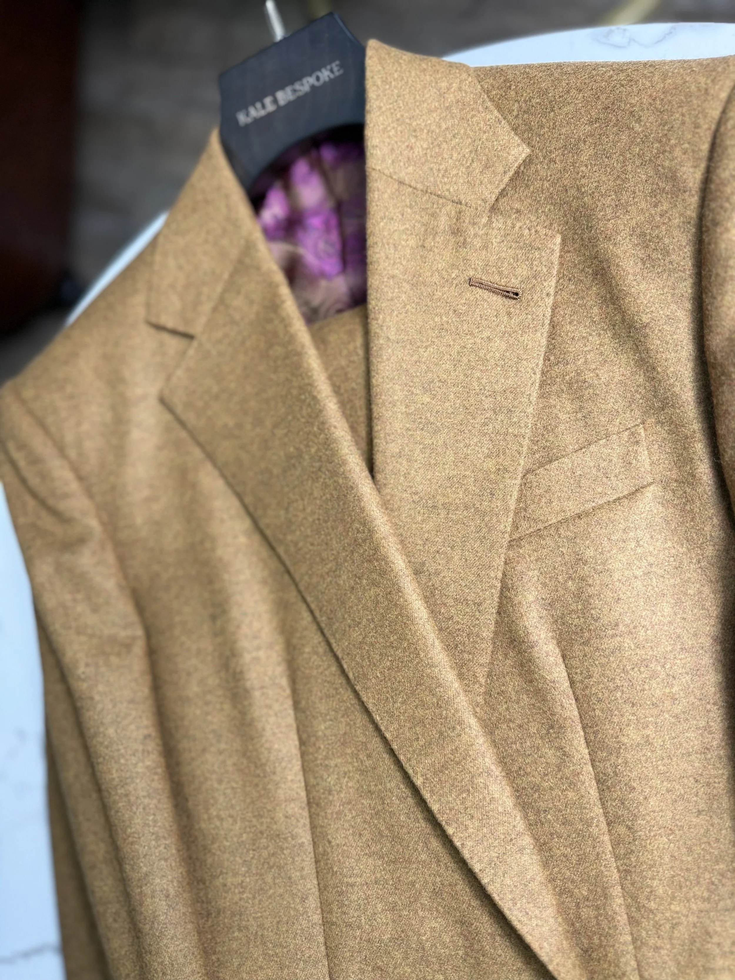 A framed mustard-colored blazer hanging on a black hanger with a purple patterned lining.