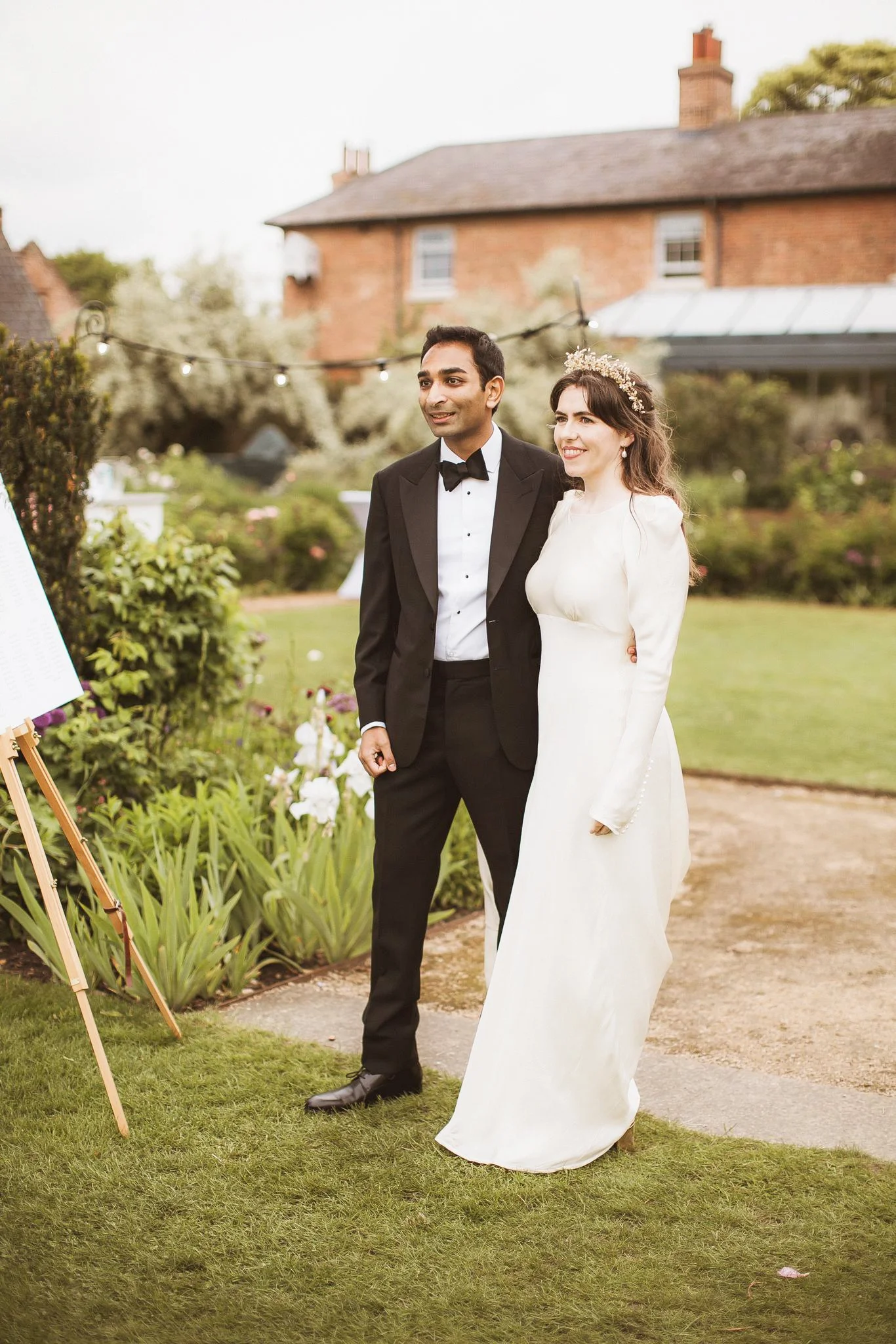 A bride and groom standing outdoors at their wedding, smiling and posing for photos. The bride is wearing a white wedding dress with long sleeves and a floral headpiece. The groom is dressed in a black tuxedo with a bow tie. The background features a