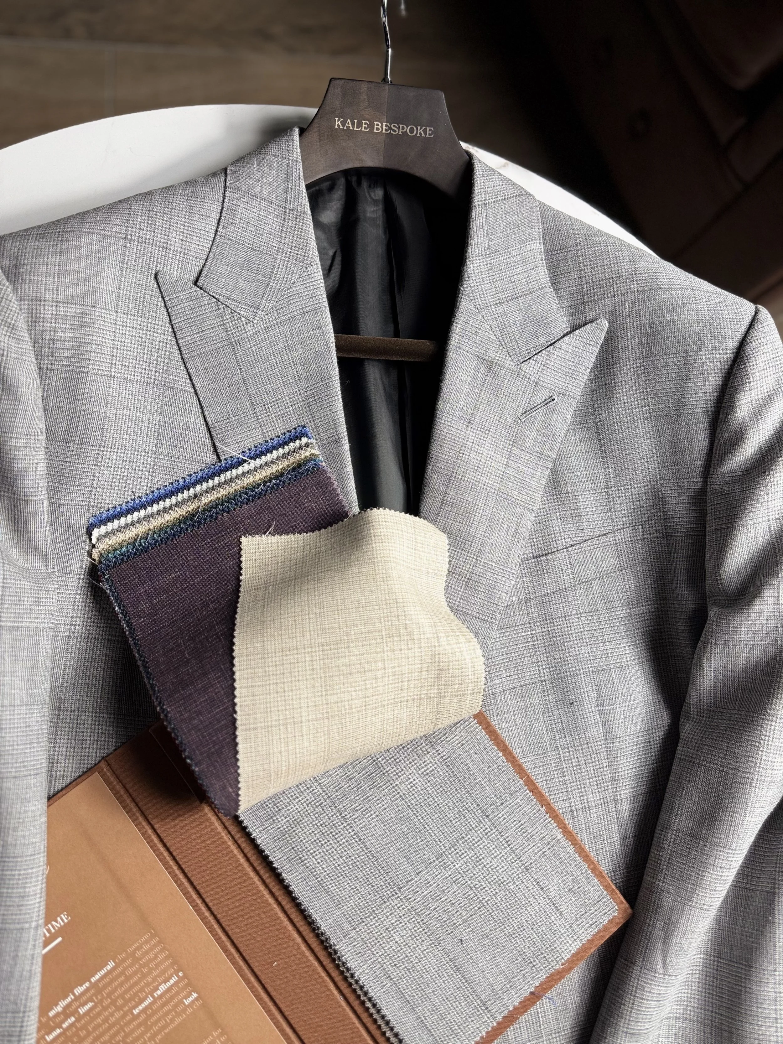 A light gray checkered blazer hanging on a wooden hanger with the label 'KALE BESPOKE'. In front of the blazer are fabric swatches in various colors and textures, along with a brown sample book.