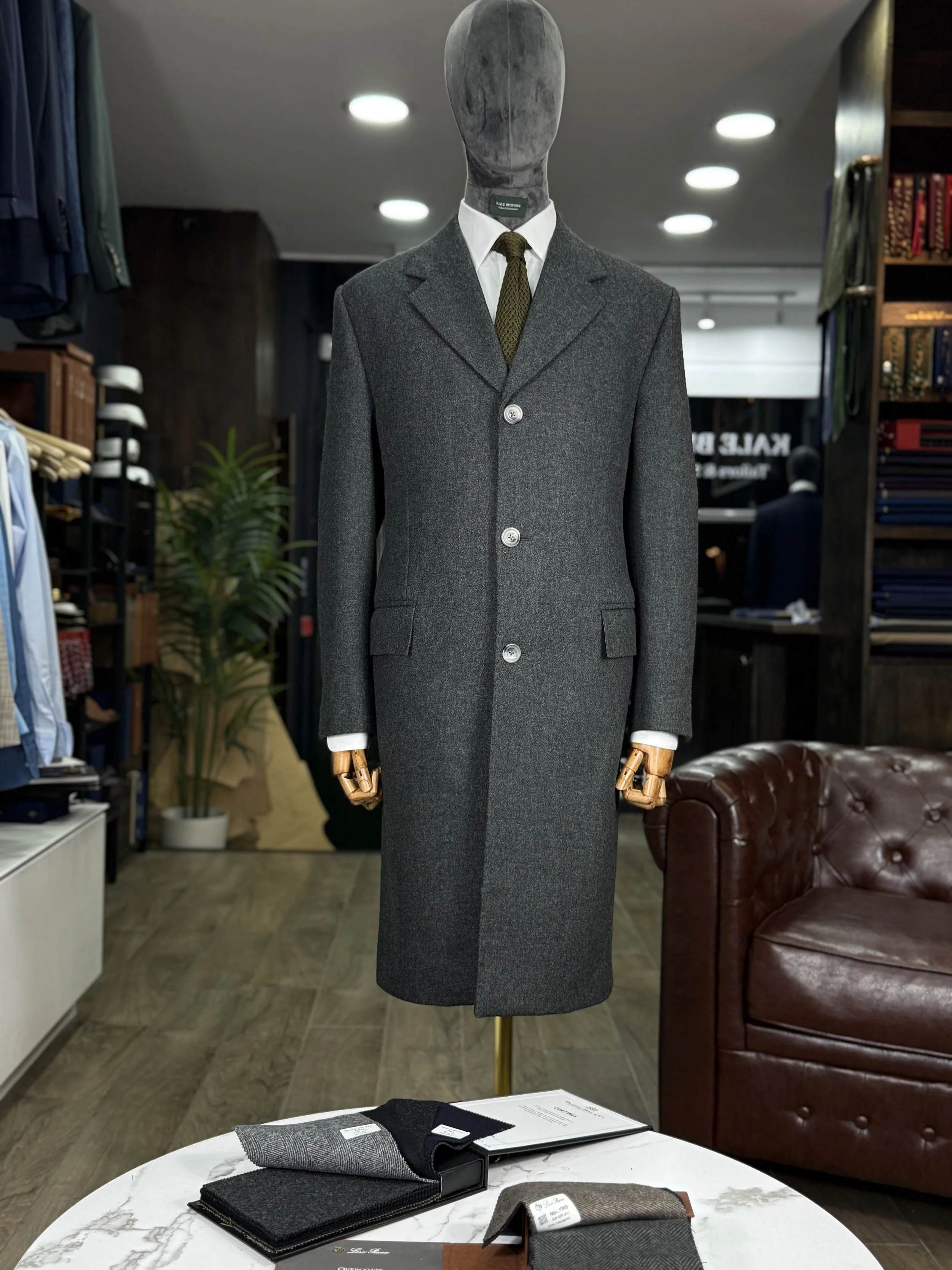 Bespoke Charcoal grey Overcaot No Chest pocket on shop mannequin with a silk knitted green silk tie.This overcaot fabric is 20oz Drim dugdale brothers full hand cut and handmade Kale bespoke Overcoat.Made by Kale Bespoke