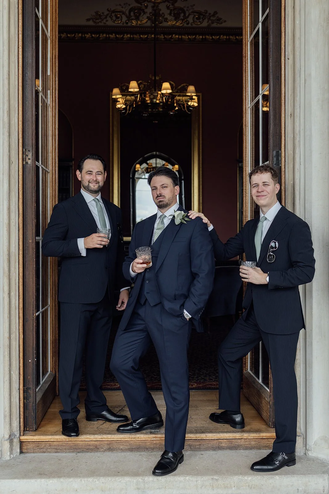 Three men in tuxedos standing in an open doorway, holding drinks at a formal event.