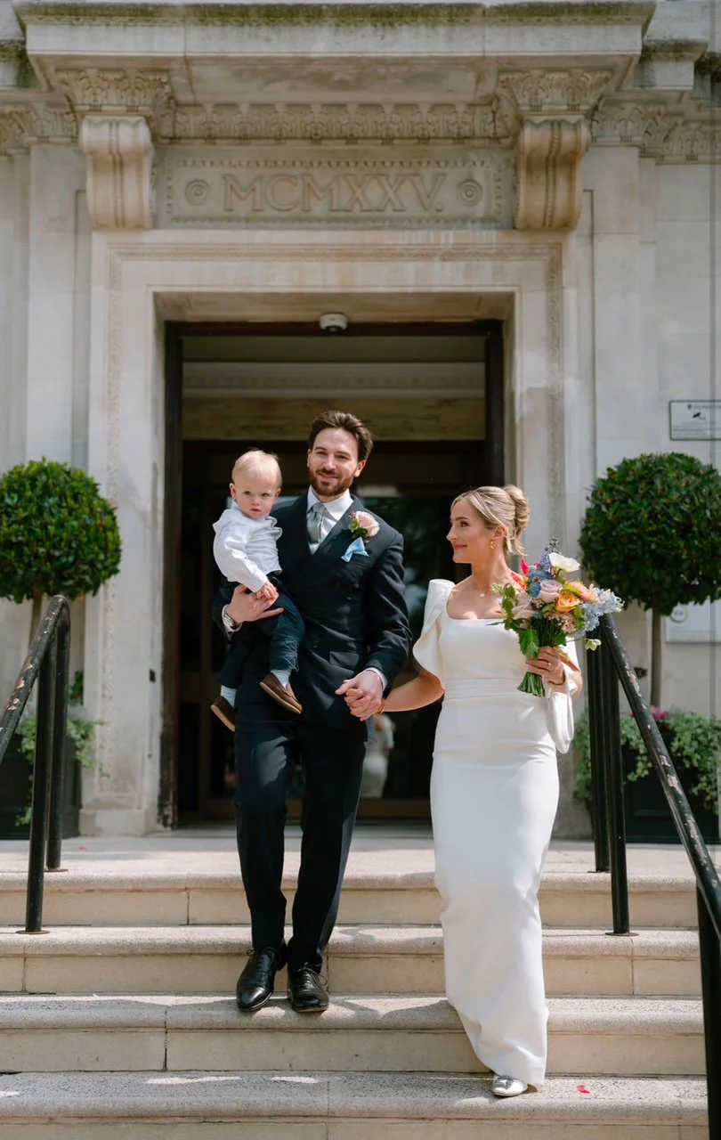 A newlywed couple and a young child descending steps from a building with an ornate stone entrance. The groom, dressed in a black suit, holds a young boy in a white shirt, while the bride, in a white wedding gown, holds a bouquet of colorful flowers.