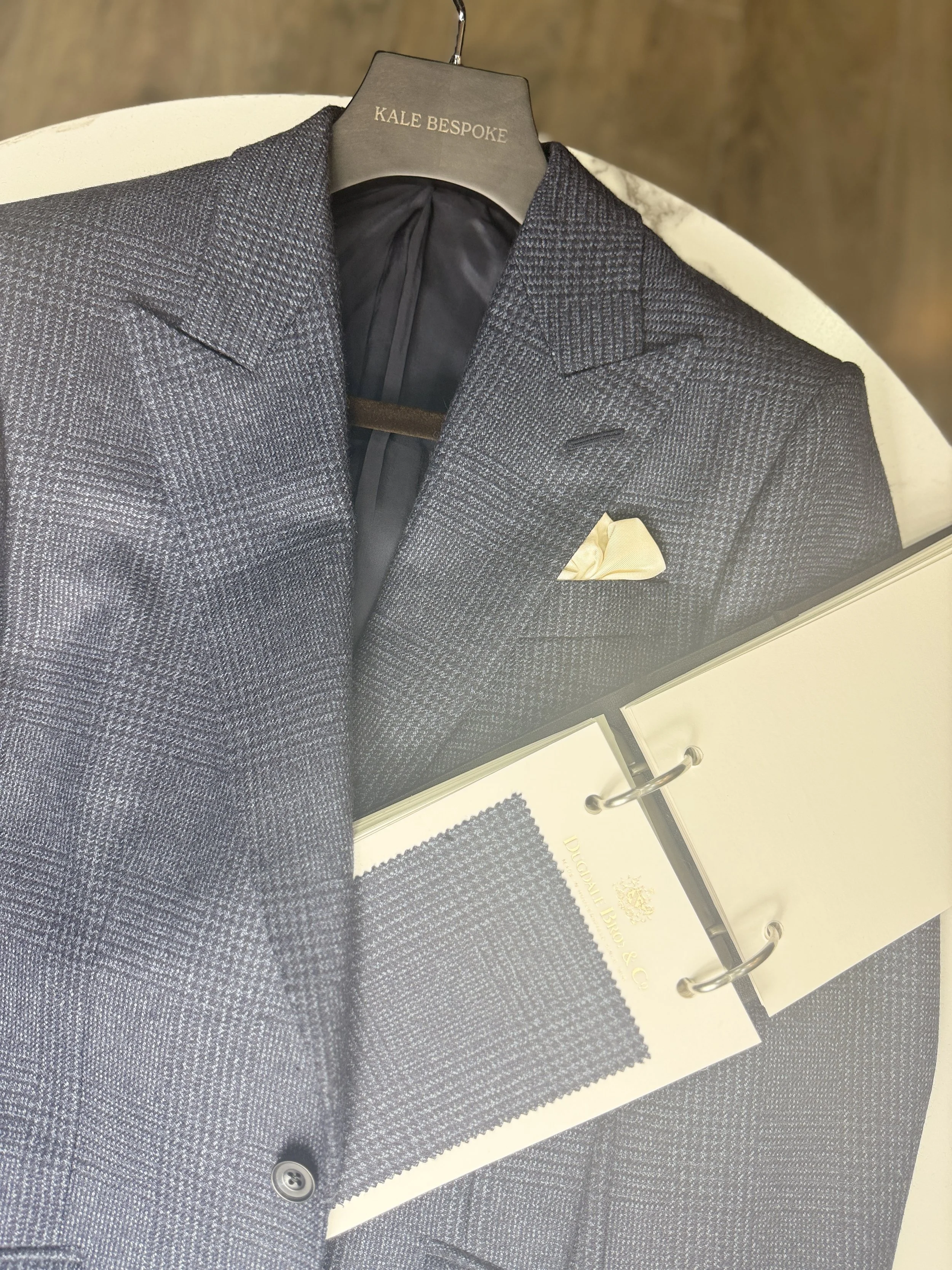 A tailored dark gray checkered blazer on a hanger with a yellow pocket square, alongside a fabric swatch with a similar pattern, and a binder with the label 'Loro Piana & C'.