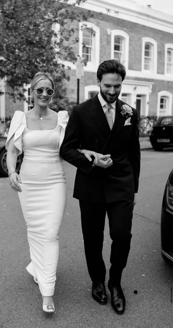 A smiling woman in a white dress and sunglasses, and a smiling man in a black suit and tie, walking arm-in-arm on a street in front of a brick building.