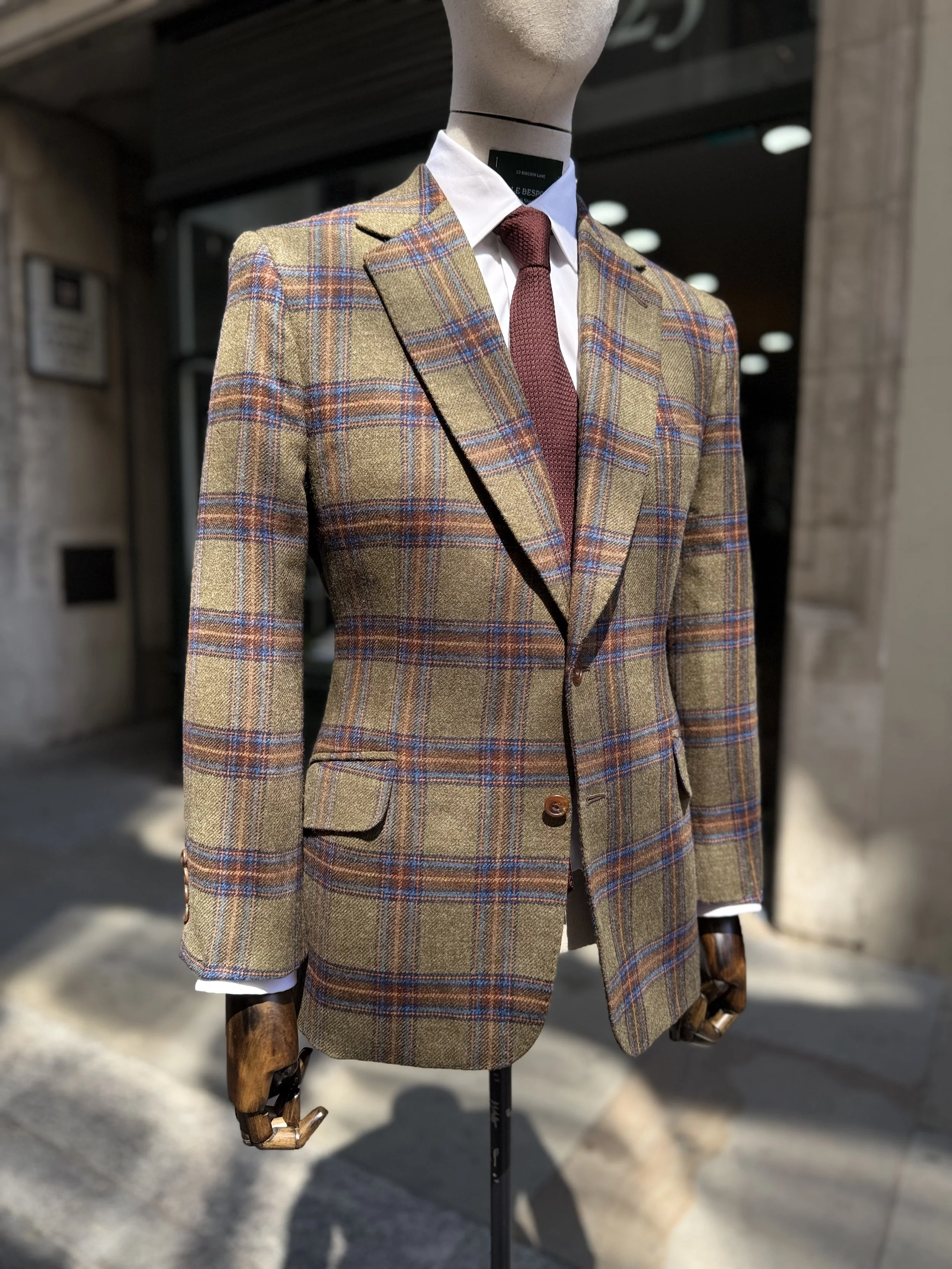 A mannequin dressed in a rally tweed fox flannel blazer, white shirt, and red grenadine tie, positioned outside a clothing store with a sign for Kale Bespoke Tailors &Shirtmakers, on  Birchin lane.