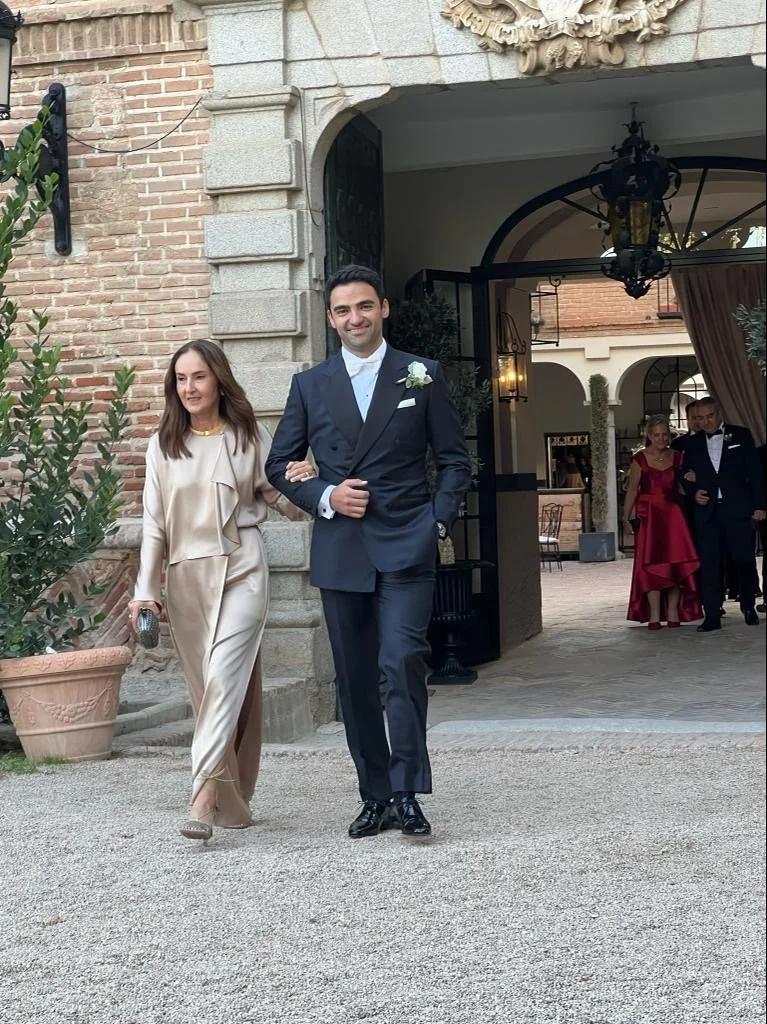 A man in a black tuxedo with a white shirt and a white boutonniere, walking arm-in-arm with a woman in an elegant beige outfit, at a formal event outside a historic building with stone and brick architecture.