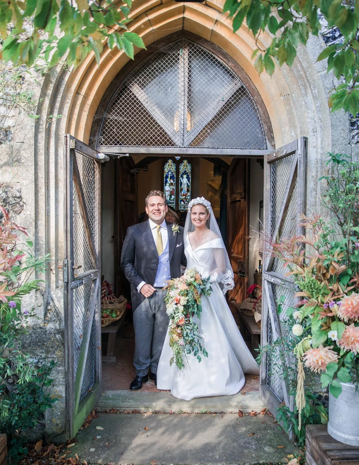 A bride and groom standing together at the entrance of a church, smiling. The bride is in a white wedding dress holding a cascading bouquet, and the groom is in a dark suit with a vest, tie, and boutonniere. The church archway is made of stone with w