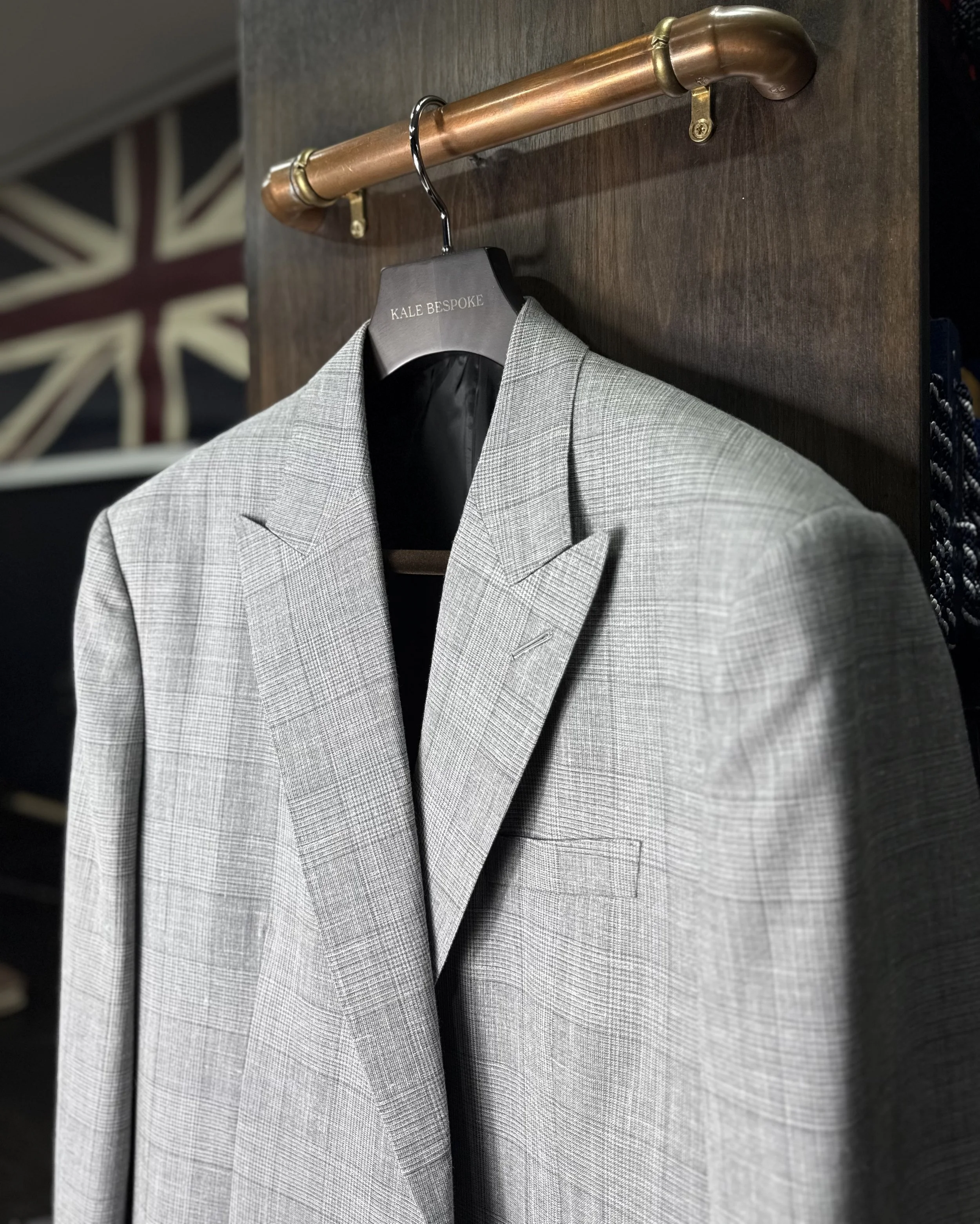A gray suit jacket with plaid pattern hanging on a black hanger labeled "KALE BESPOKE" on a wooden rack. In the background, there's a part of a wall with a Union Jack flag.