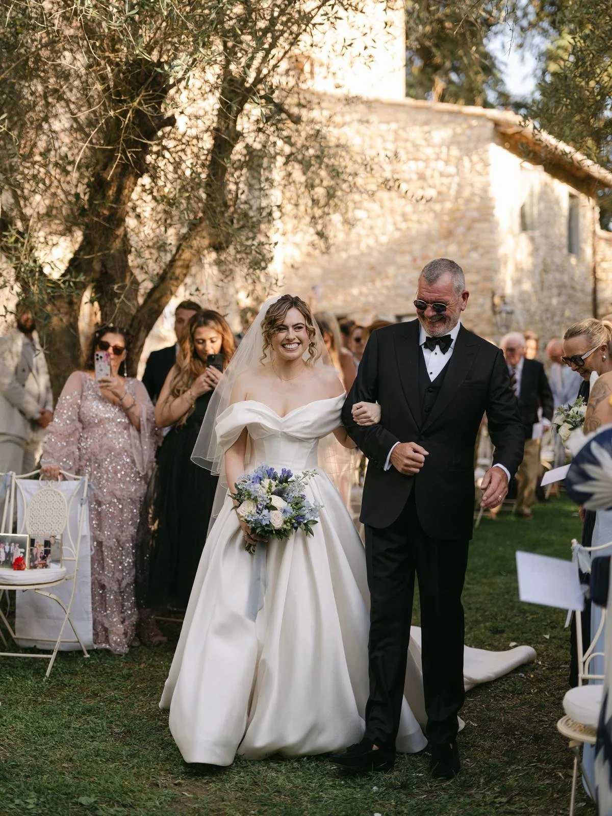 A bride in a white wedding gown holding a bouquet and smiling, walking down the aisle with an older man in a black tuxedo, at an outdoor wedding ceremony surrounded by guests.