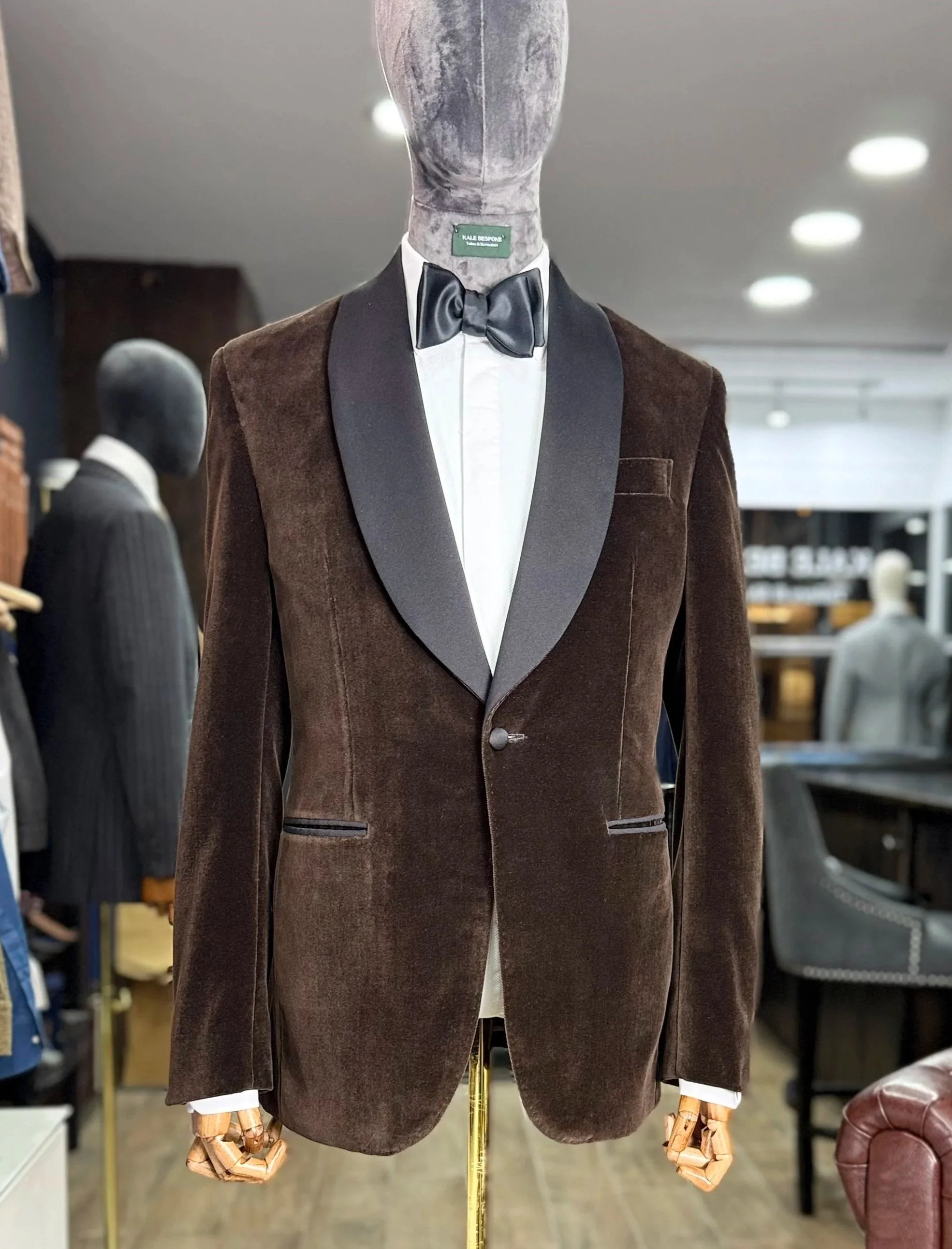 Mannequin wearing a dark brown velvet tuxedo jacket with black satin lapels, a white dress shirt, and a black bow tie inside a clothing store.