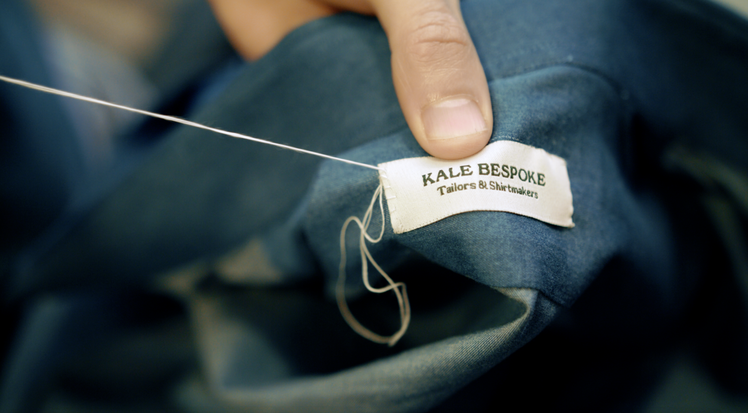 Close-up of a clothing label reading 'KALE BESPOKE Tailors & Shirtmakers' on a teal fabric, with a hand holding the fabric and a sewing needle attached.