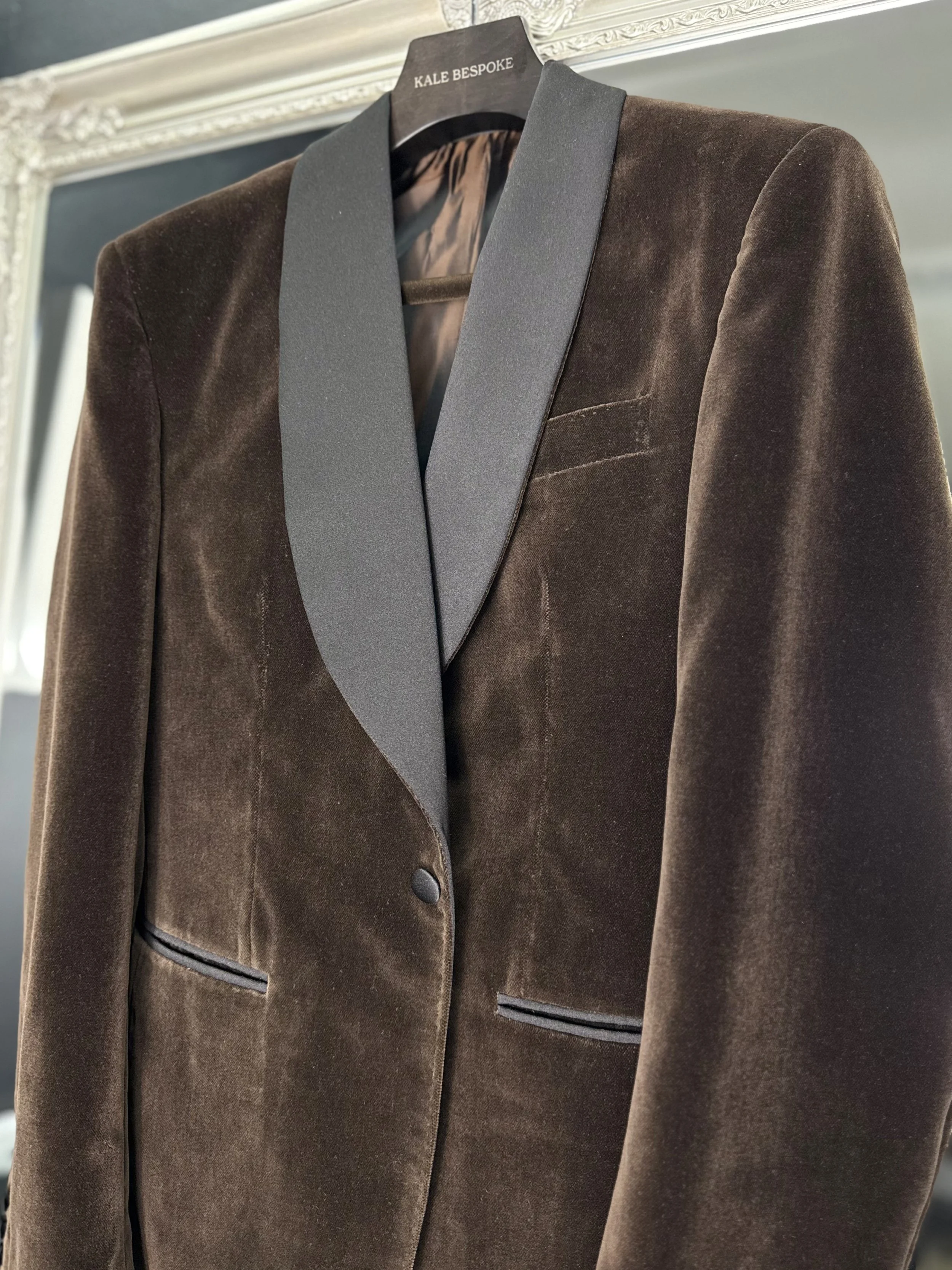 A brown velvet blazer with black satin lapels hanging on a black hanger labeled 'KALE BESPOKE', with intricate white molding on the wall behind.
