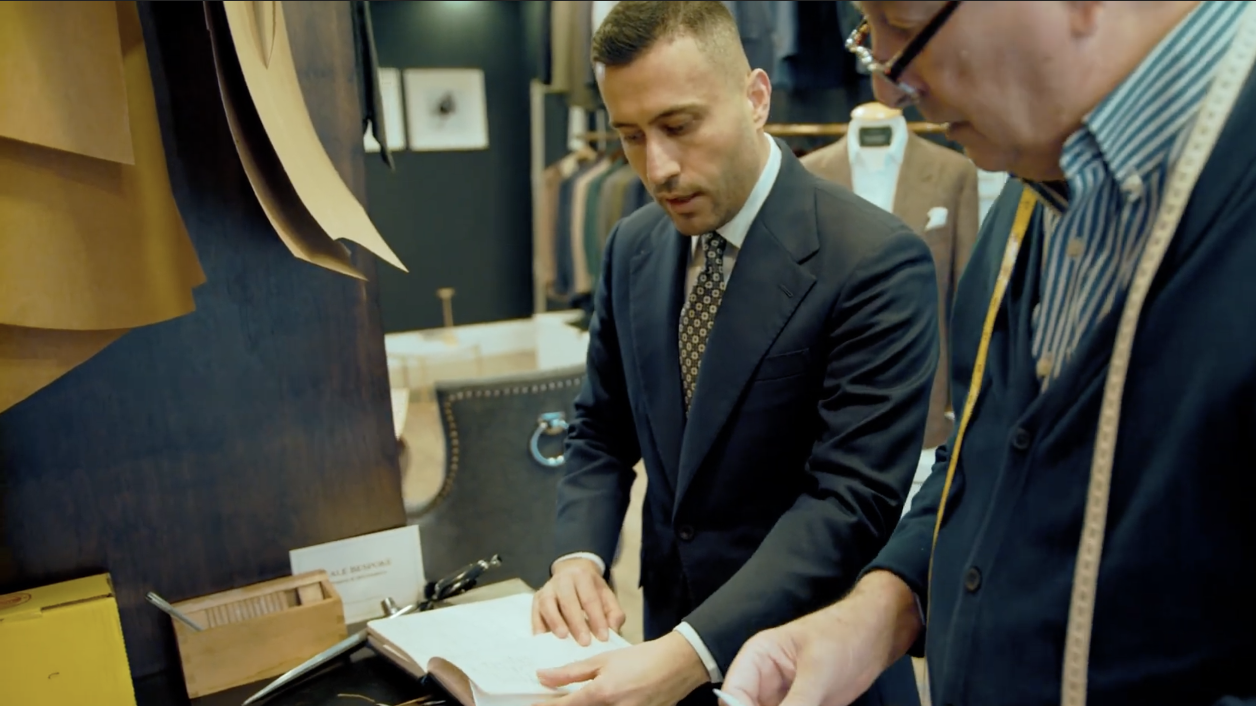 Two men in formal suits looking at a book in a clothing store.