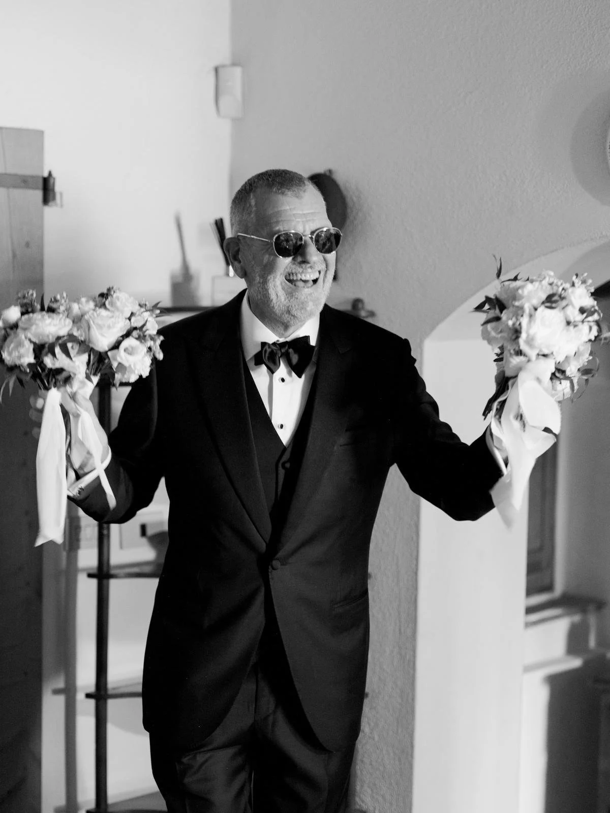 A man in a tuxedo and sunglasses smiling and holding a bouquet of flowers in each hand.Kale bespoke three piece tuxedo for big day