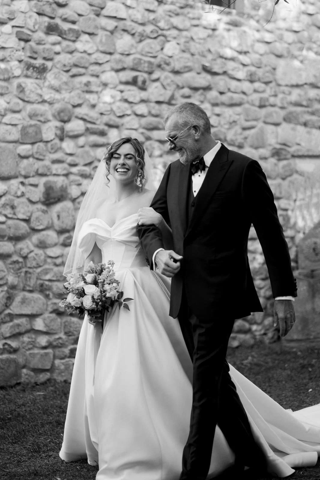 A bride in a wedding dress holding a bouquet walking arm-in-arm with an older man in a tuxedo near a stone wall, smiling and looking happy.