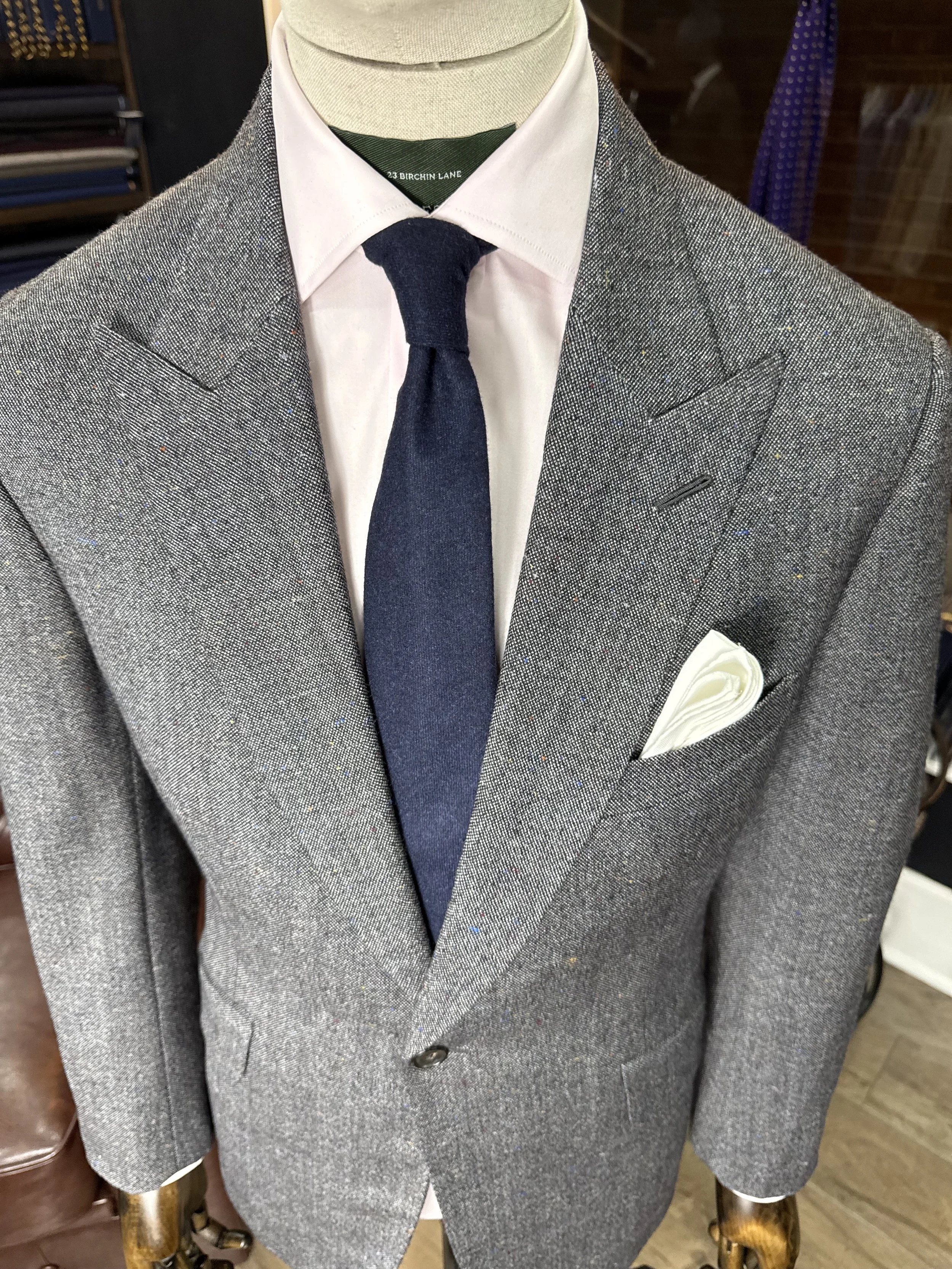 Mens grey patterned suit jacket, white dress shirt, dark blue tie, and white pocket square on a mannequin.