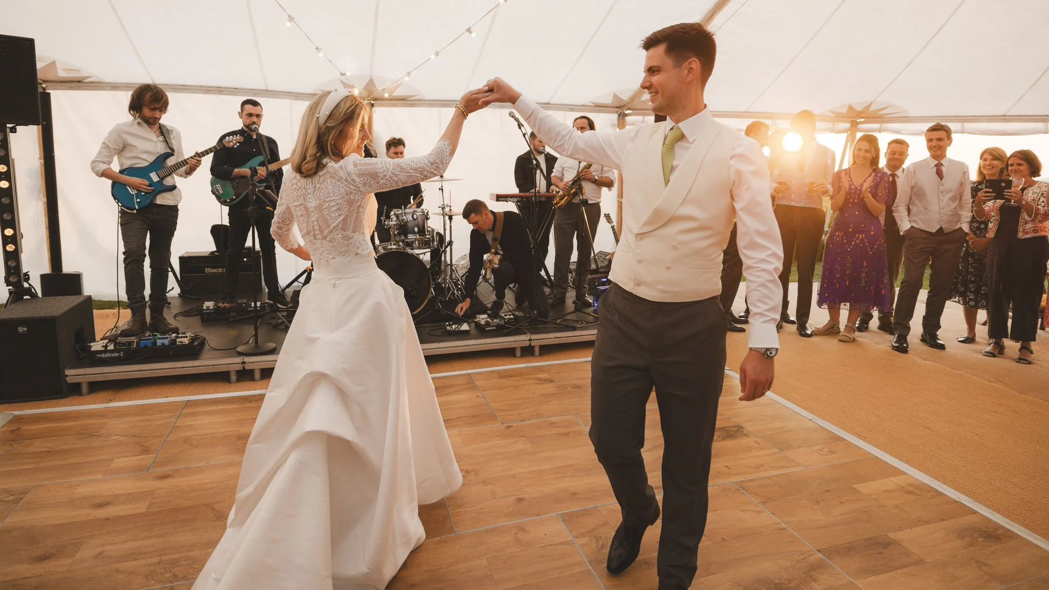 A bride and groom are dancing together during a wedding reception, with a live band playing on stage in the background and guests watching and taking photos.