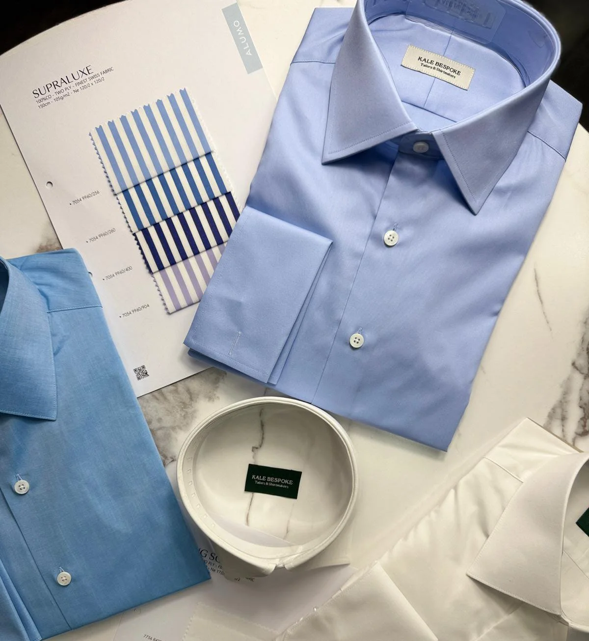 Flat lay of light blue and white dress shirts, fabric swatches with blue stripes, and a roll of white fabric on a marble surface, for tailoring or fashion design.