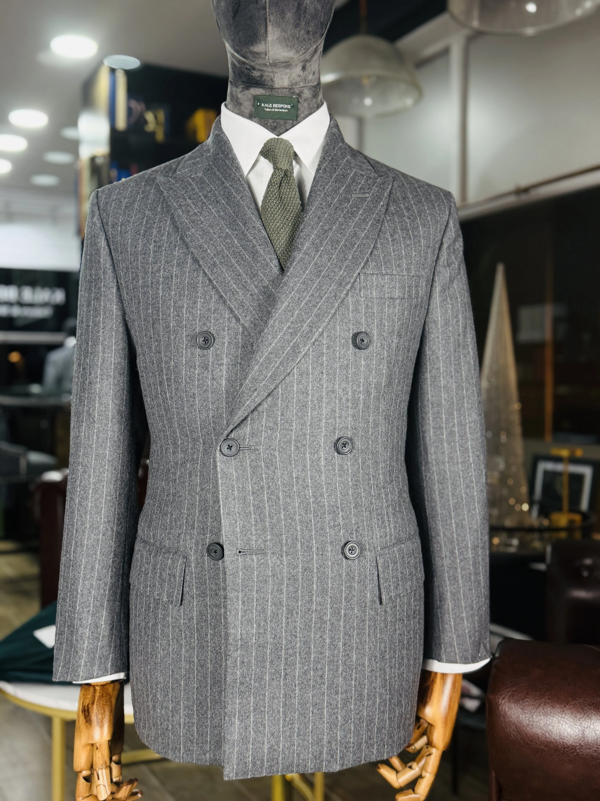 A mannequin dressed in a gray double-breasted plaid suit with a white shirt and an olive green knit tie, positioned in a retail store with blurred background shelves and lighting.