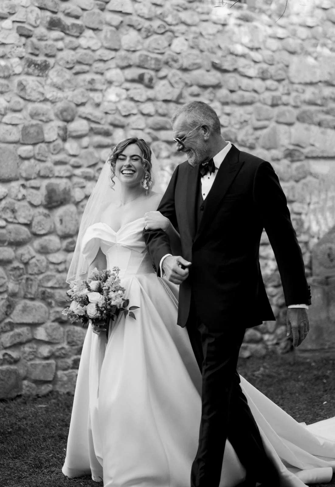A bride in a wedding dress holding a bouquet of flowers is smiling as she walks arm-in-arm with a groom in a tuxedo, against a stone wall.