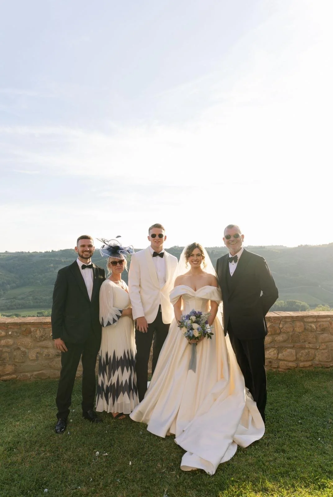 A wedding party with five people standing outdoors on a grassy area near a stone wall, with rolling hills in the background under a clear sky. The bride is in the center wearing an off-the-shoulder white wedding gown, holding a bouquet of flowers. To