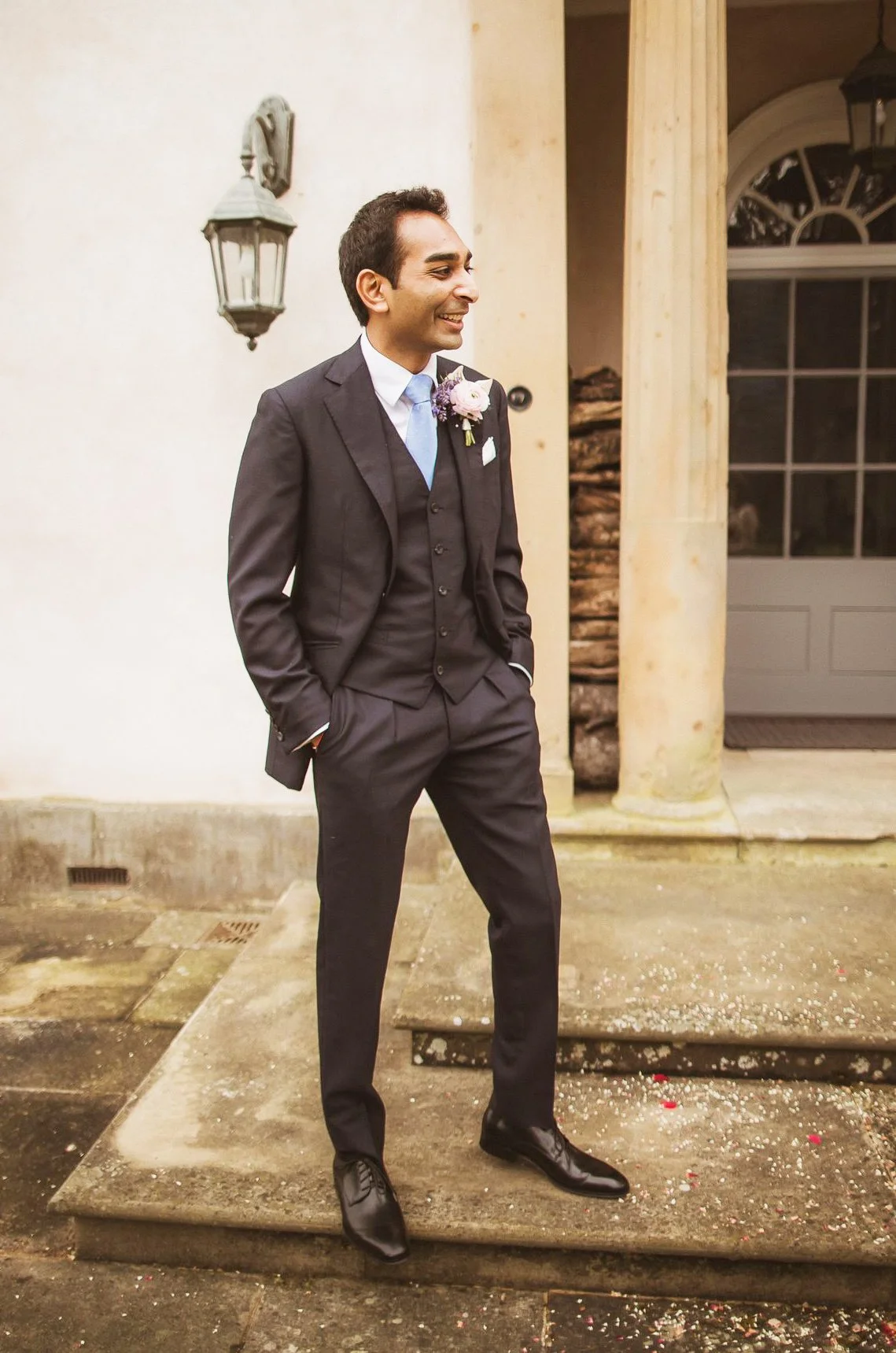 A man in a black suit with a light blue tie and a pink flower boutonniere is standing outside a building, smiling, with his hands in his pockets.