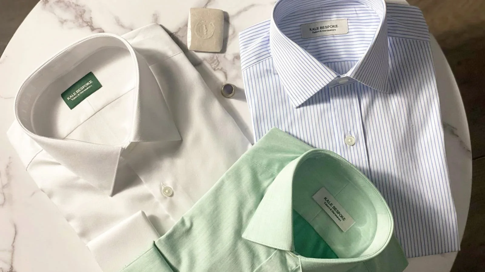 Three folded dress shirts in white, light green, and blue with stripes, displayed on a marble surface, with a small soap and a ring beside them.