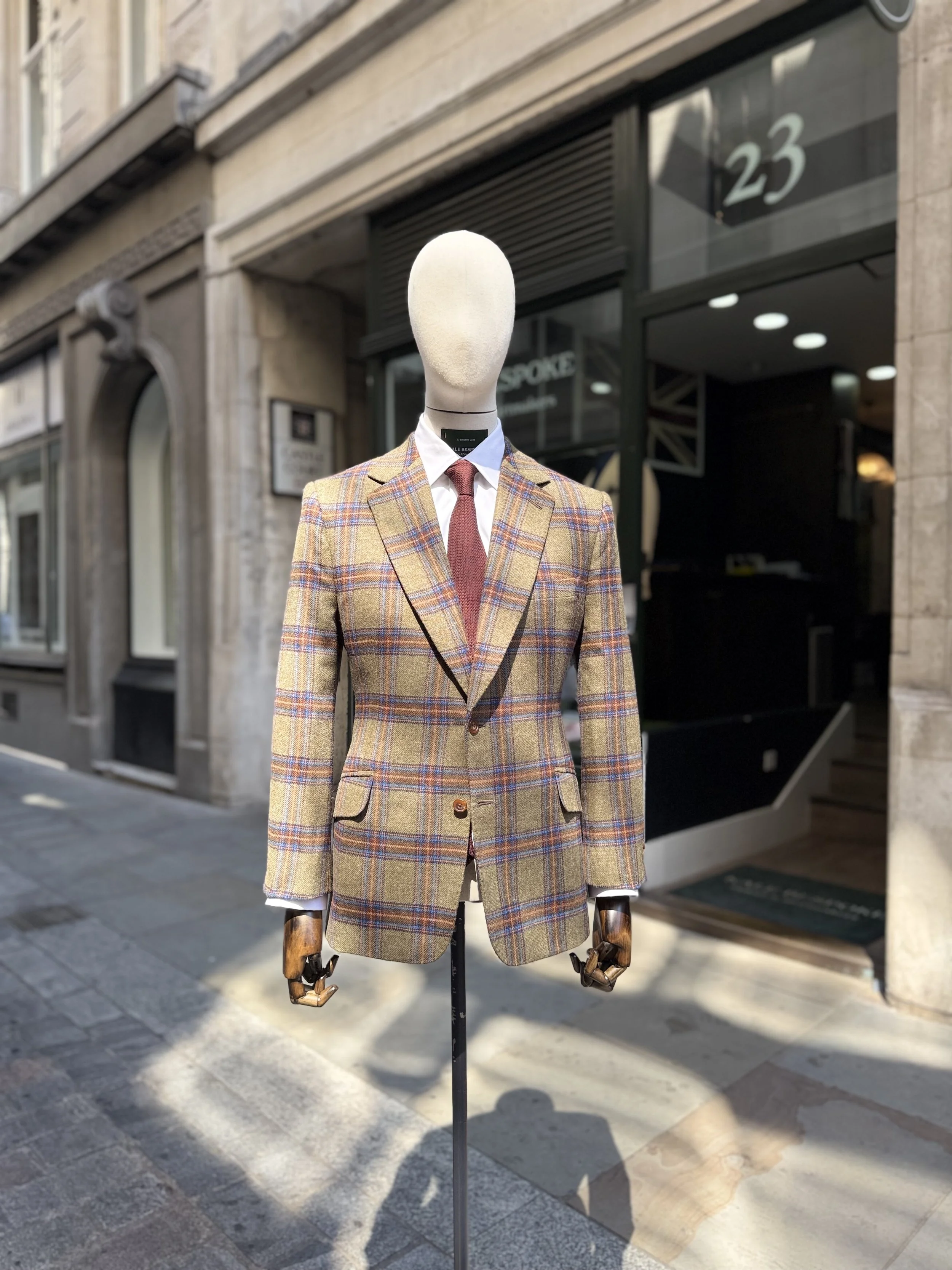 A mannequin dressed in a rally tweed fox flannel blazer, white shirt, and red grenadine tie, positioned outside a clothing store with a sign for Kale Bespoke Tailors &Shirtmakers, on  Birchin lane.
