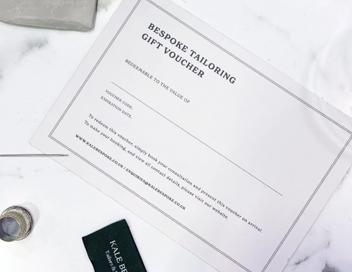 A bespoke tailoring gift voucher lying on a white surface, with a black fabric tag and a small bottle nearby.