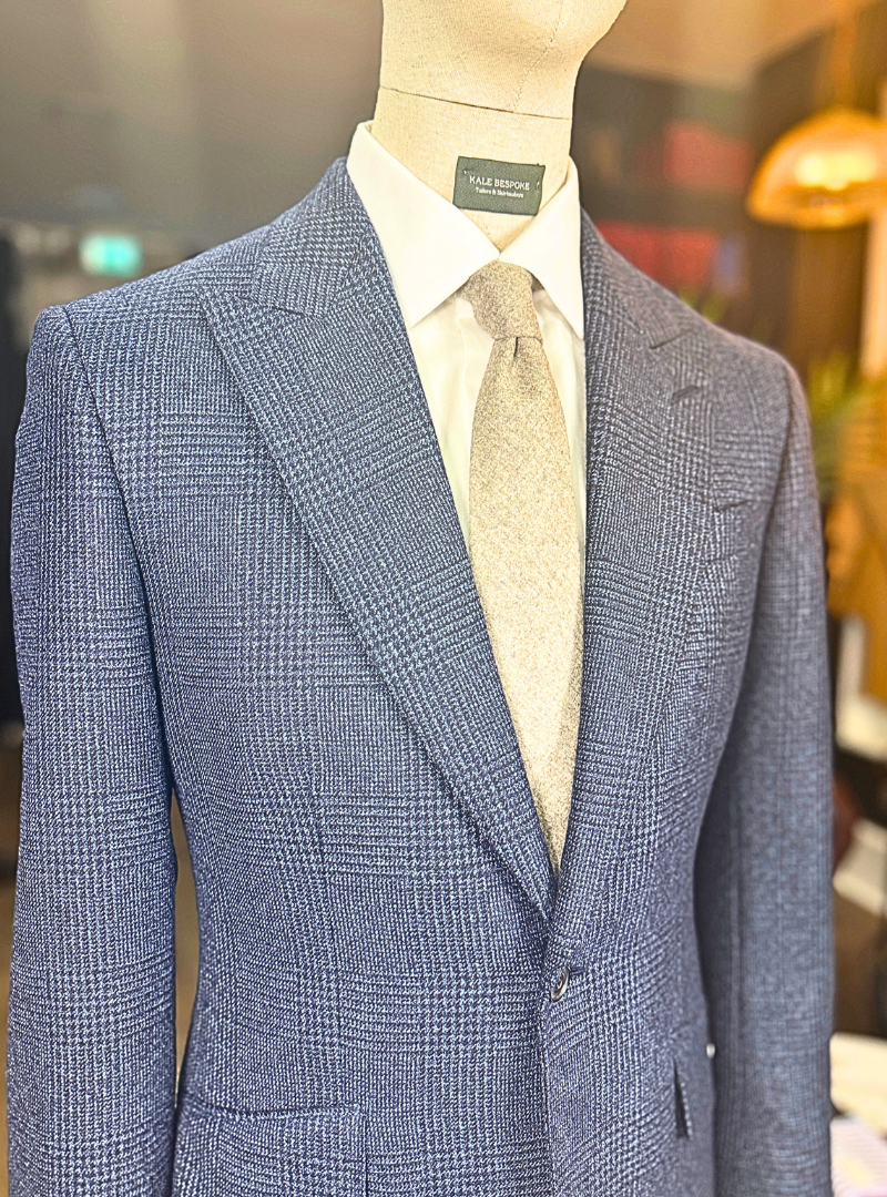 Close-up of a mannequin dressed in a blue plaid suit jacket, white collared shirt, and light gray tie, with a blurred background of a retail store.