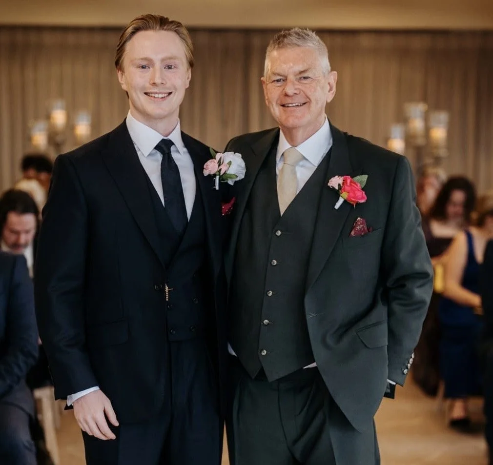Groom and father wearing bespoke three
piece wedding suits by Kale Bespoke, London. Classic black and deep green tailored suits with waistcoats, silk ties, and boutonnières, photographed during an indoor wedding ceremony. Handmade bespoke tailoring 
