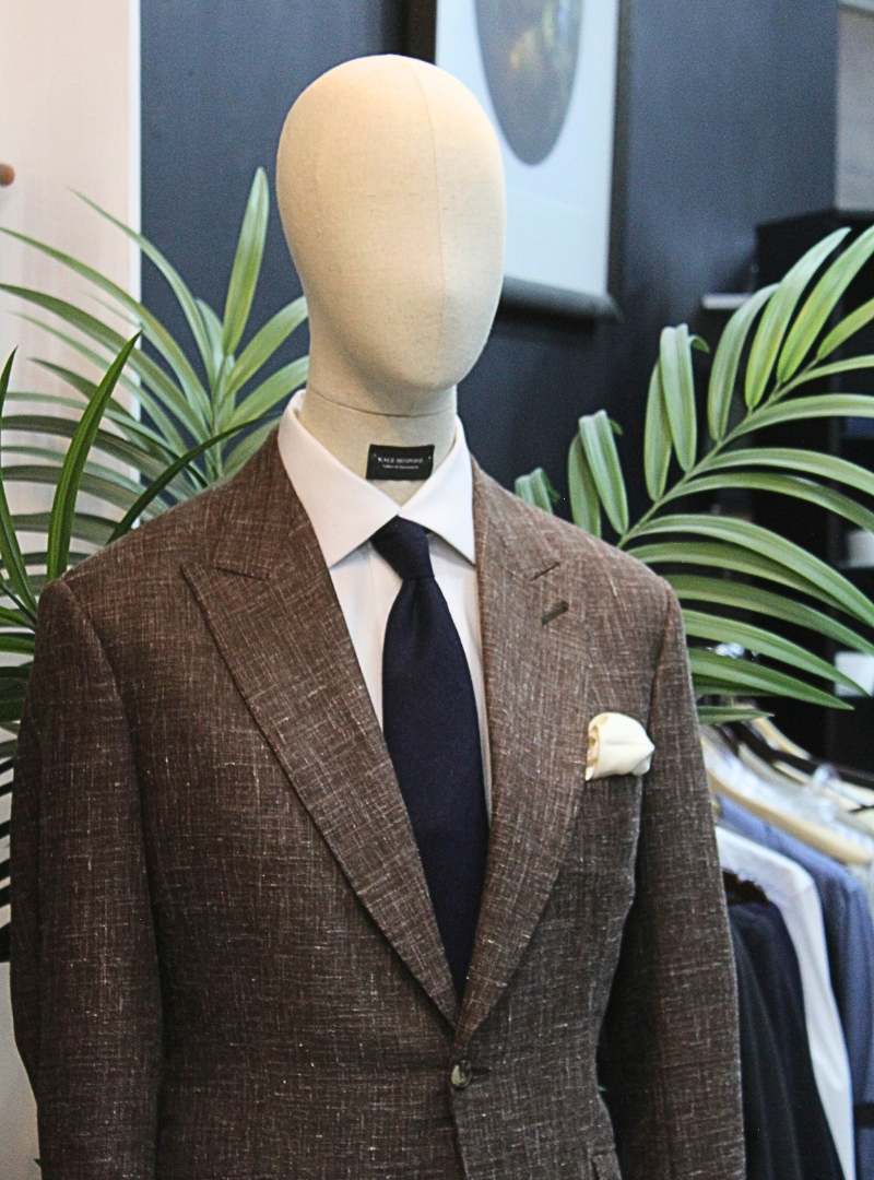 A mannequin dressed in a brown textured blazer, white dress shirt, navy tie, and white pocket square, standing in front of green plants and a black background.
