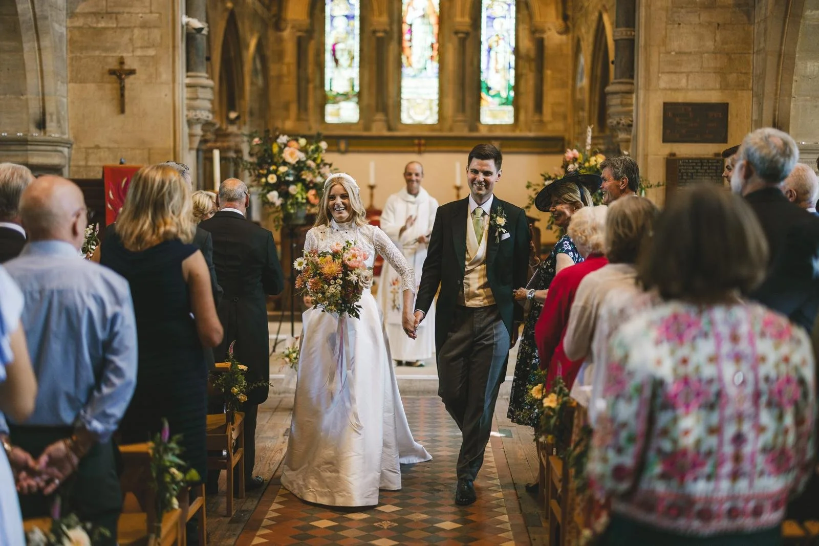 A newly married couple walks down the aisle holding hands inside a church, surrounded by family and friends, after their wedding ceremony.