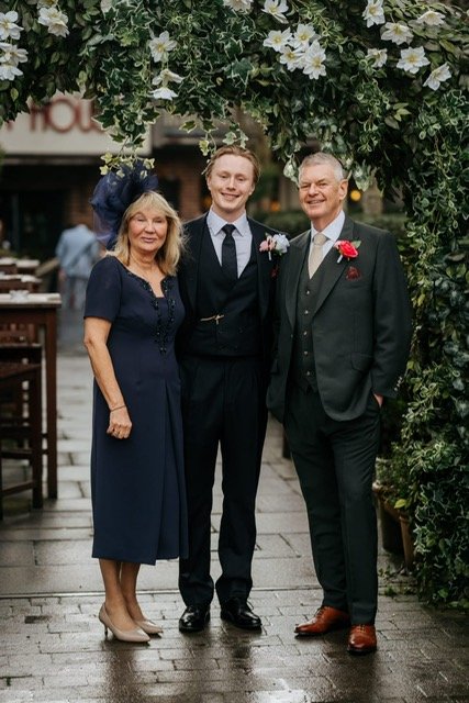 Groom with parents wearing bespoke three-piece wedding suits by Kale Bespoke, London. Classic black and deep green tailored suits featuring waistcoats, silk ties, and boutonnières, photographed outdoors under floral arch at a wedding venue. Handmade 